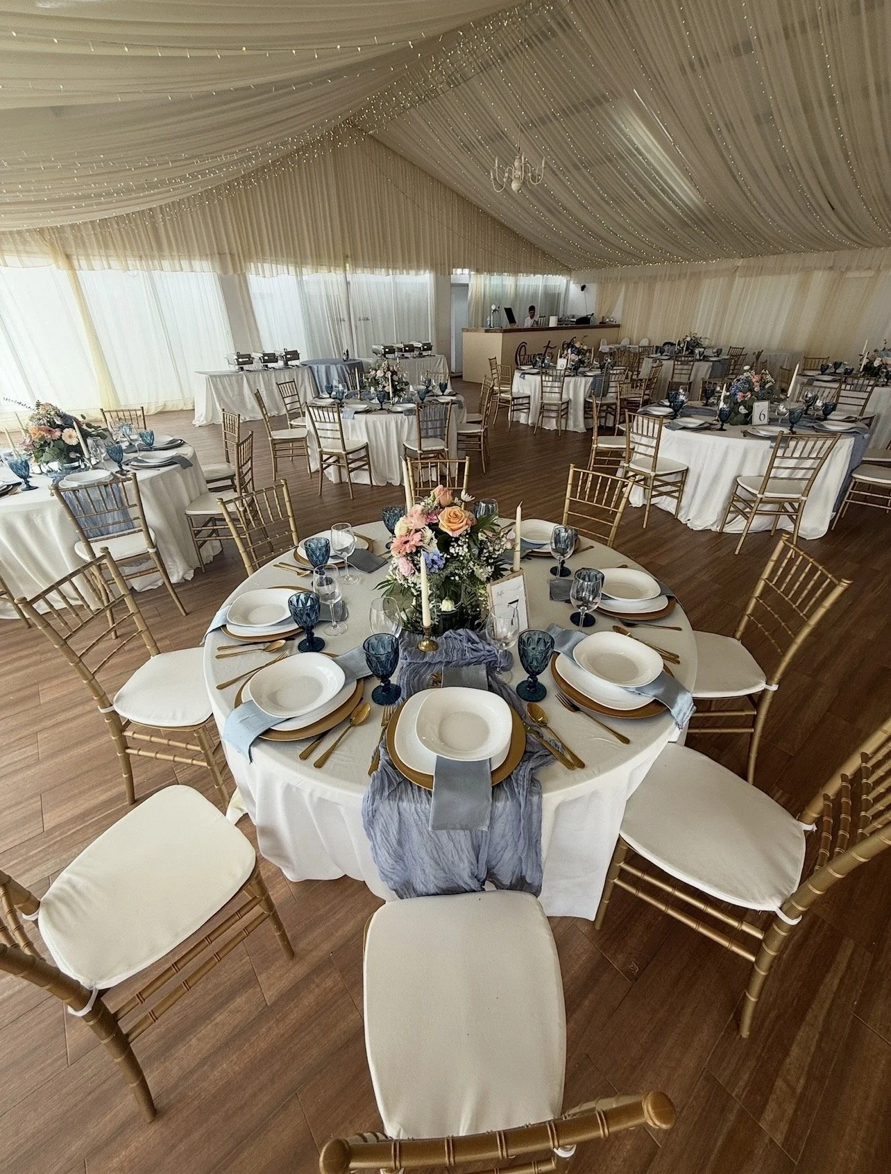 Elegant banquet hall decorated for a wedding reception with round tables, gold chairs, white tablecloths, floral centerpieces, and draped ceiling with fairy lights.