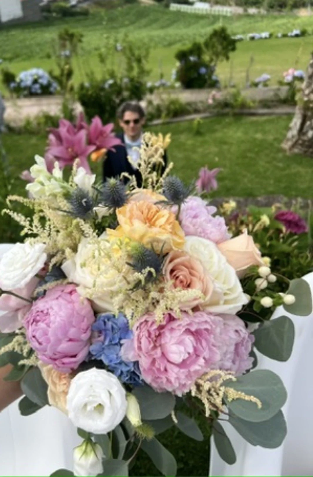 Close-up of a colorful flower bouquet with pink peonies, roses, and other mixed flowers, with a person wearing sunglasses and a dark jacket in the background in a garden setting.