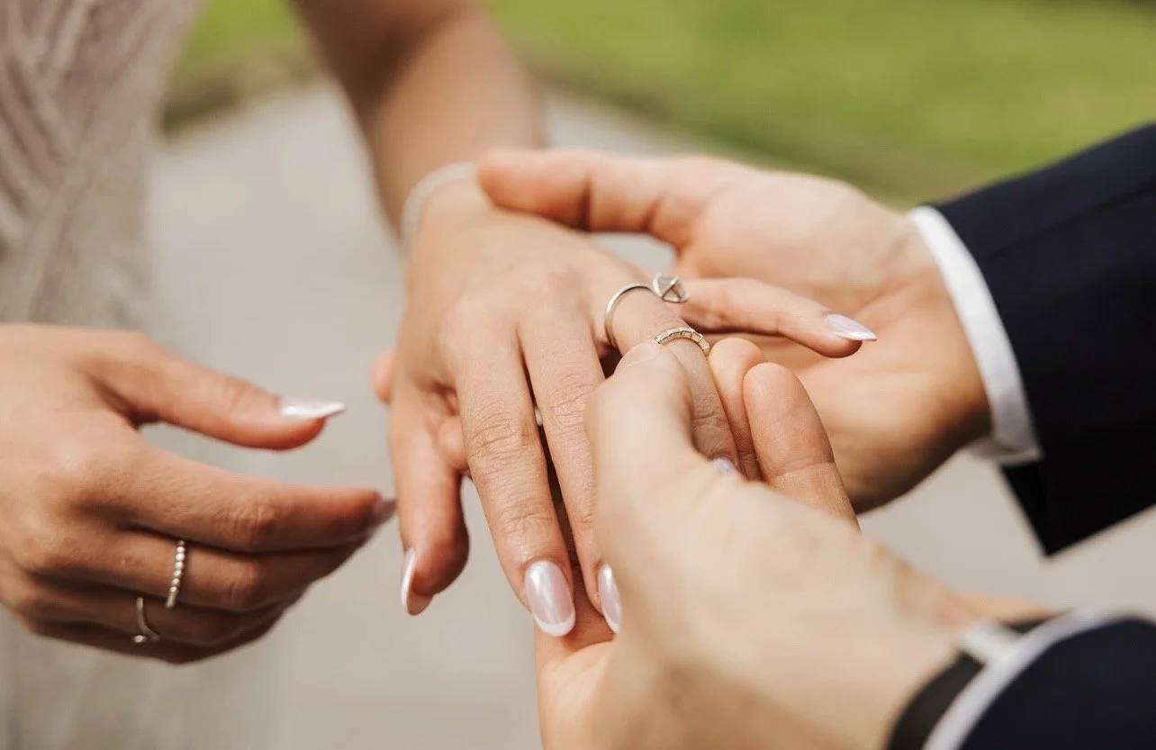 Couple exchanging wedding rings outdoors, with close-up focus on hands and rings.