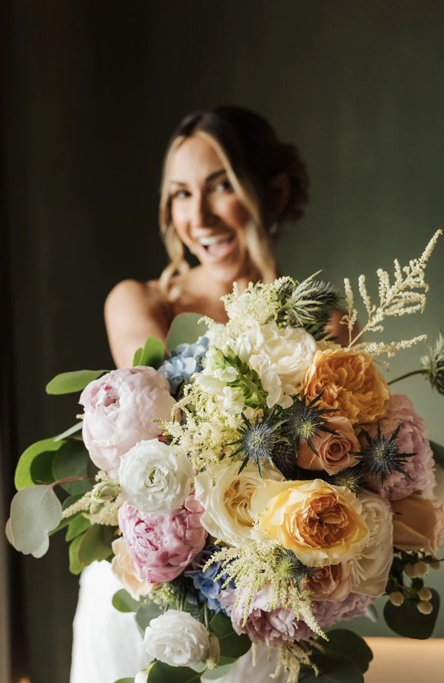 A smiling woman holding a large, colorful bouquet of flowers, including roses, peonies, and greenery, with a blurred background.