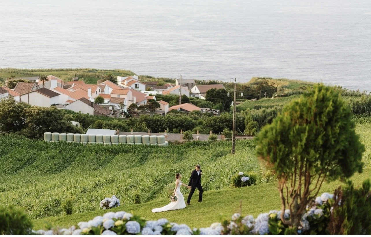 A bride and groom walking hand in hand on a grassy hill, with a village and the ocean in the background.
