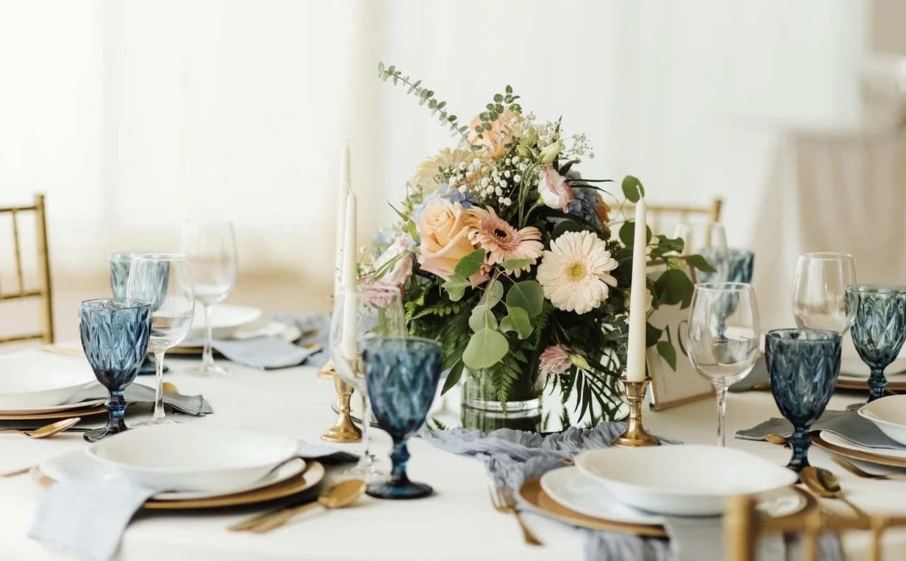 Elegant table setting with a floral centerpiece, white plates, gold utensils, blue and clear glasses, and white taper candles, arranged for a formal event.