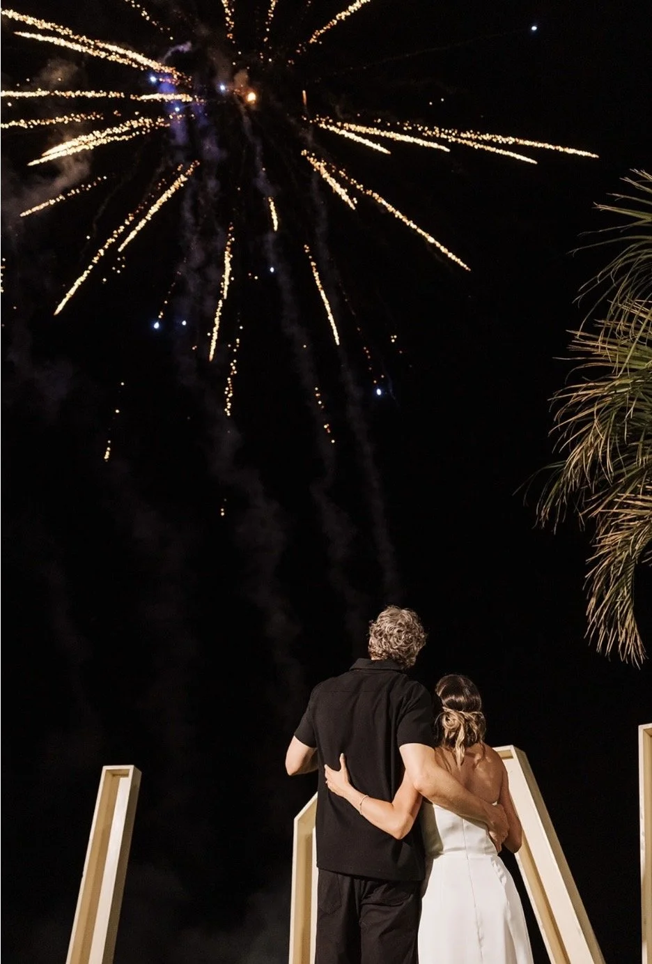 A couple watching fireworks at night, standing close together with the man's arm around the woman's waist. The fireworks are bright and colorful against the dark sky, with some palm trees visible on the right side.