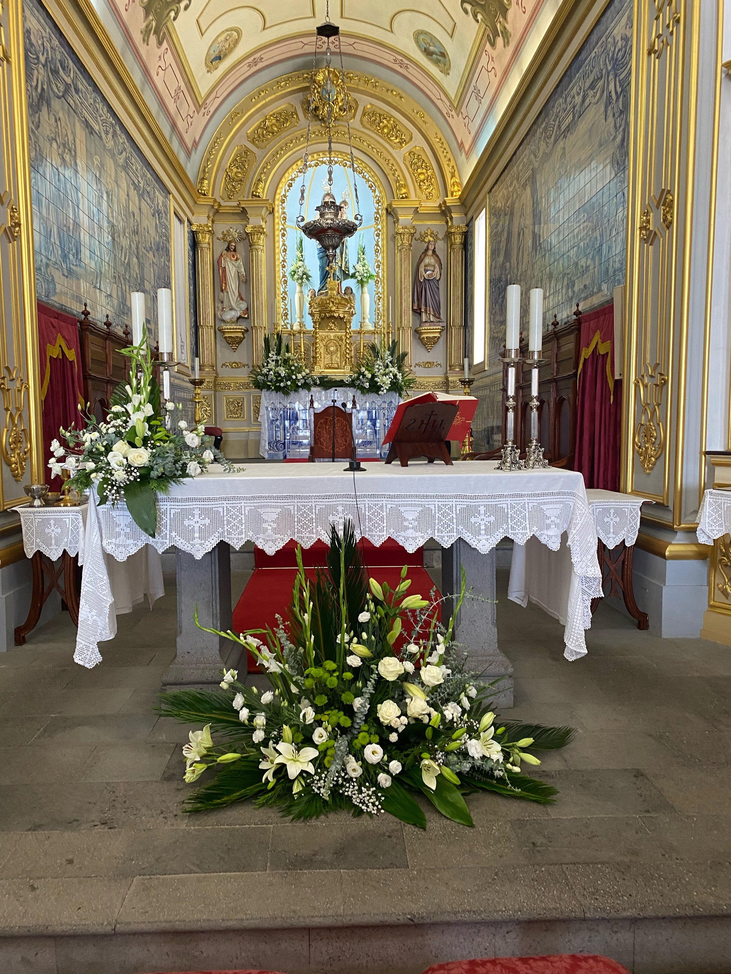 Interior of a church with an altar decorated with white flowers, candles, and religious statues, ornate gold and white detailing, and a large floral arrangement in front.