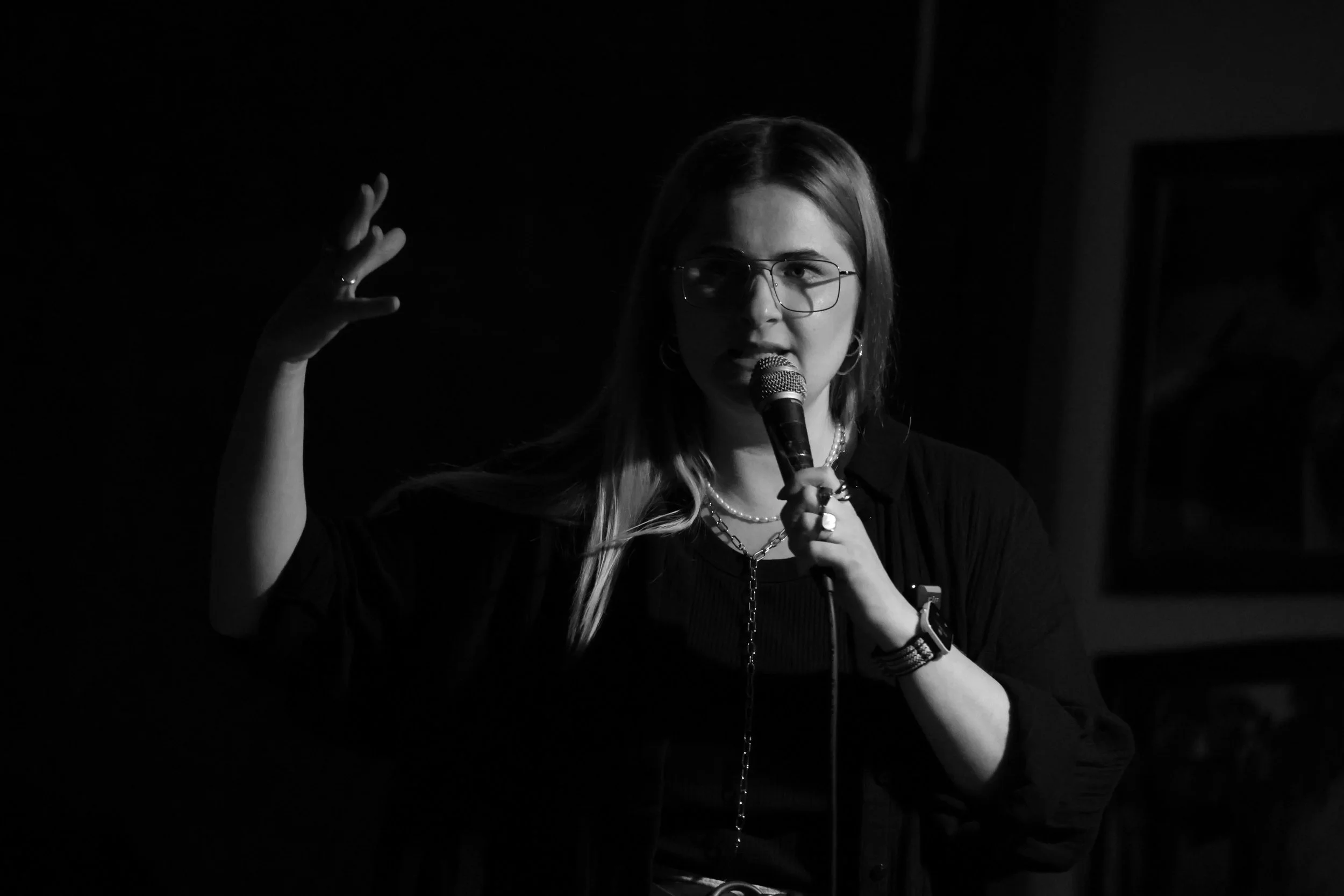 Alina Van Riva hält ein Mikrofon in der Hand, das Bild ist schwarz-weiß, sie hält eine Hand nach oben als würde sie mit kreisenden Bewegungen auf ihren Kopf deuten