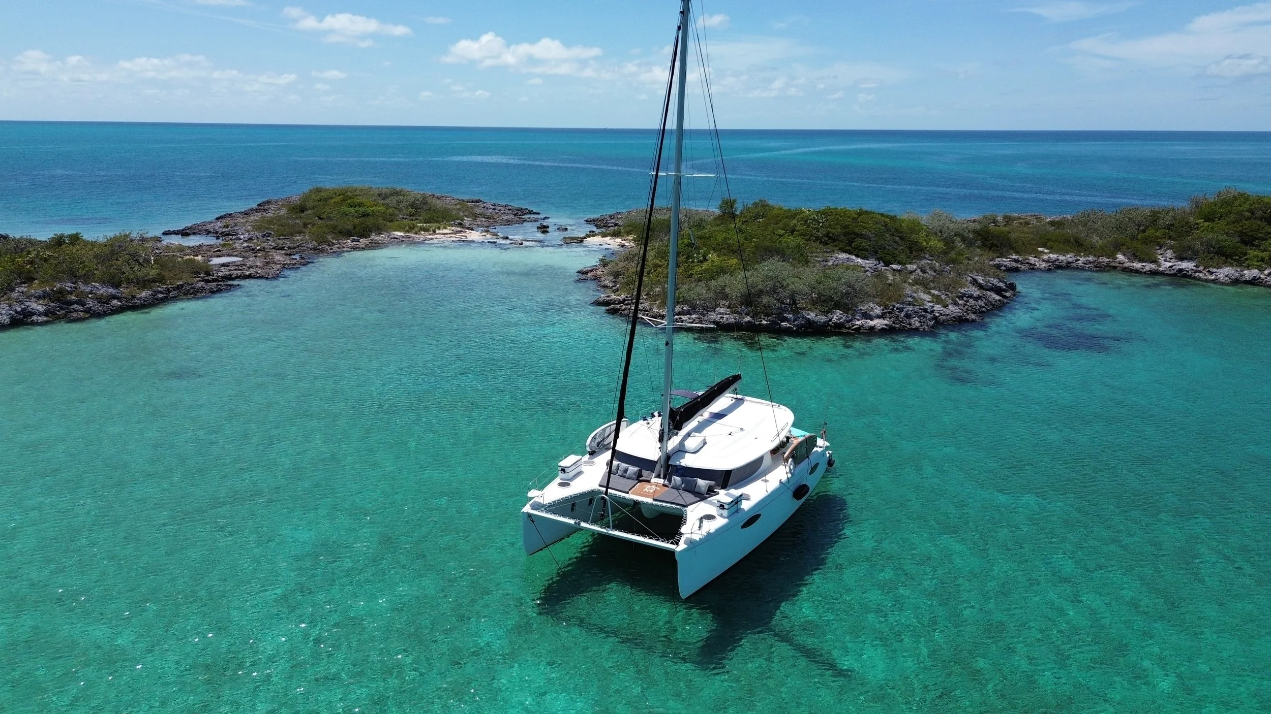A white sailing yacht anchored in clear turquoise water near a small island with rocky shores and green vegetation, with the open ocean and partly cloudy sky in the background.