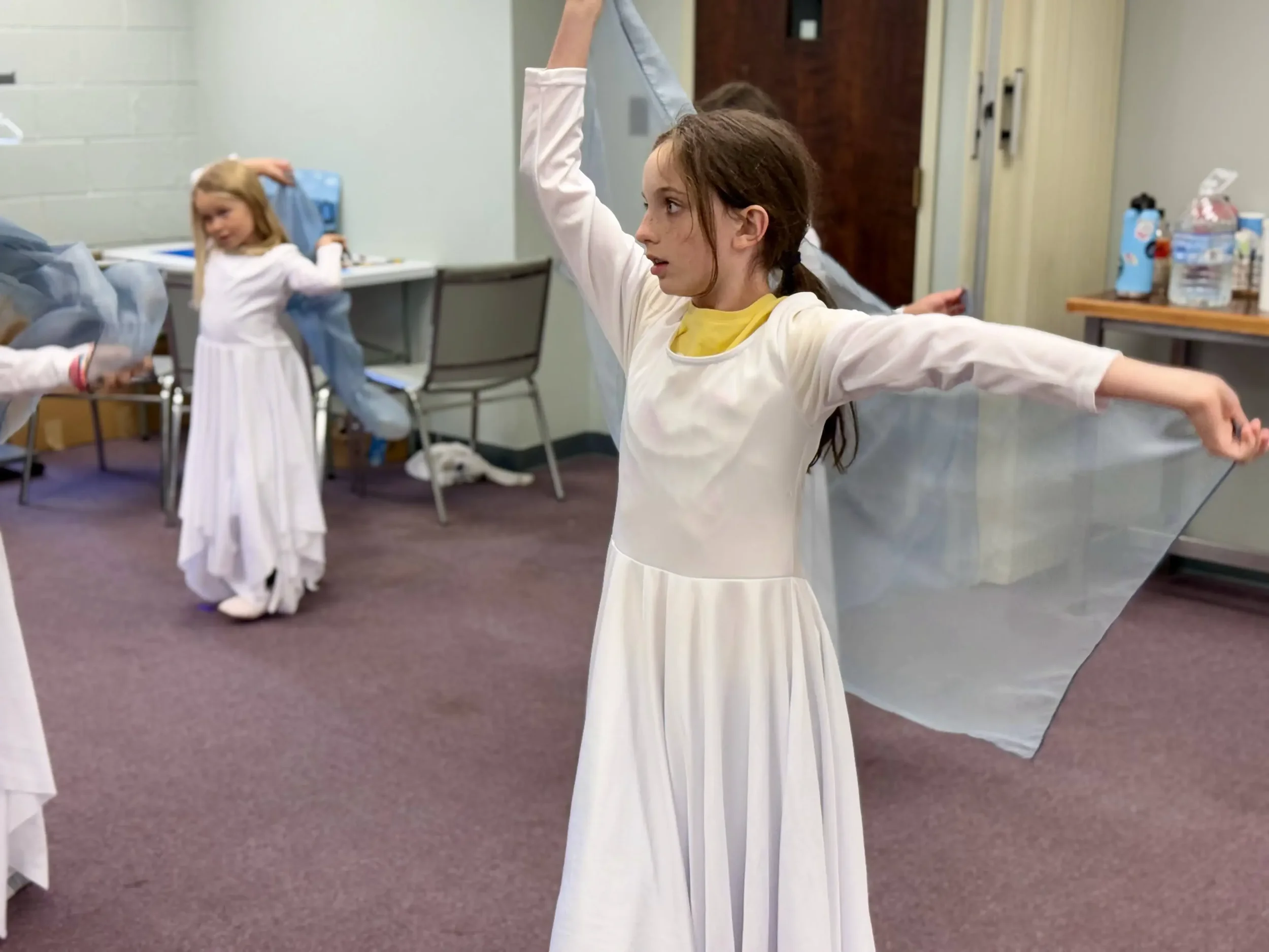  Young dancers in white dresses rehearse worship dance movements with flowing blue scarves. 