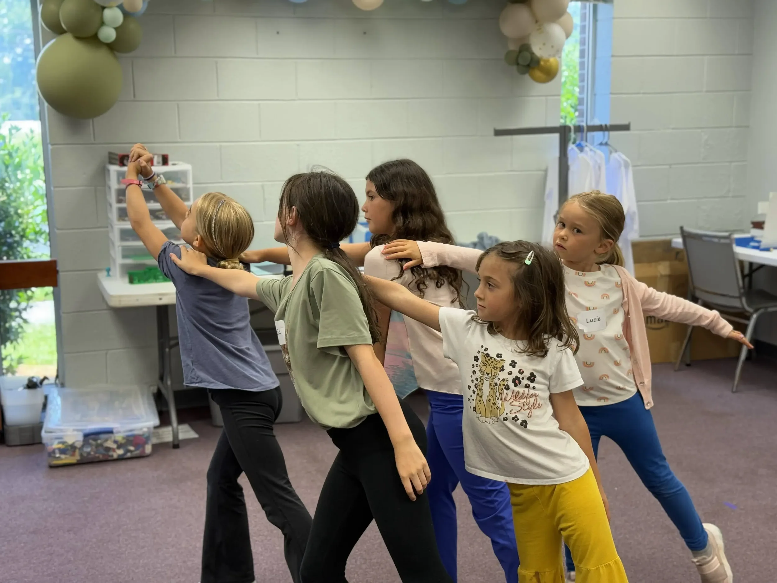  A group of children practice coordinated dance movements with hands on each other’s shoulders. 