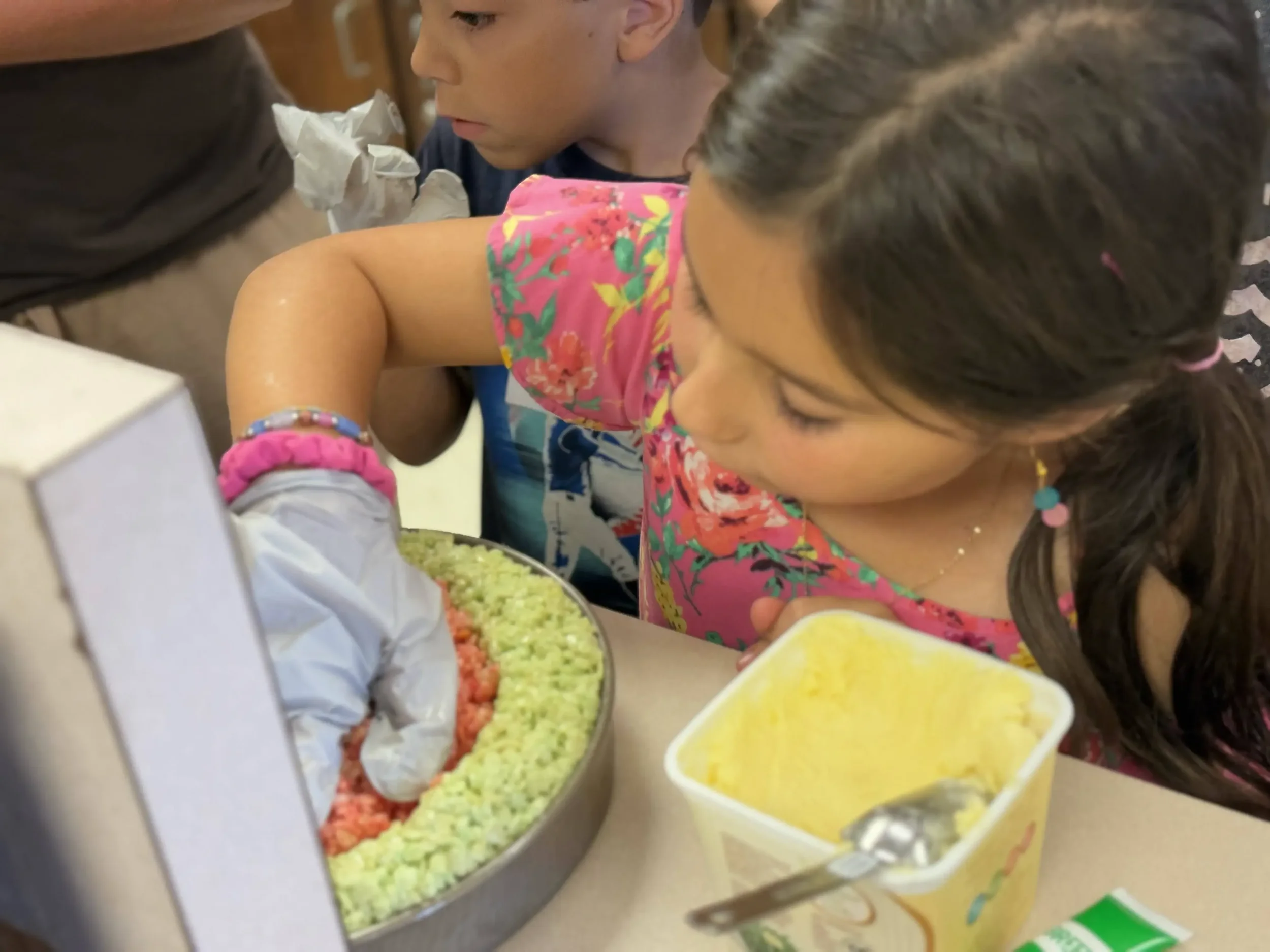  Children press colorful cereal layers into a round pan while preparing a dessert. 