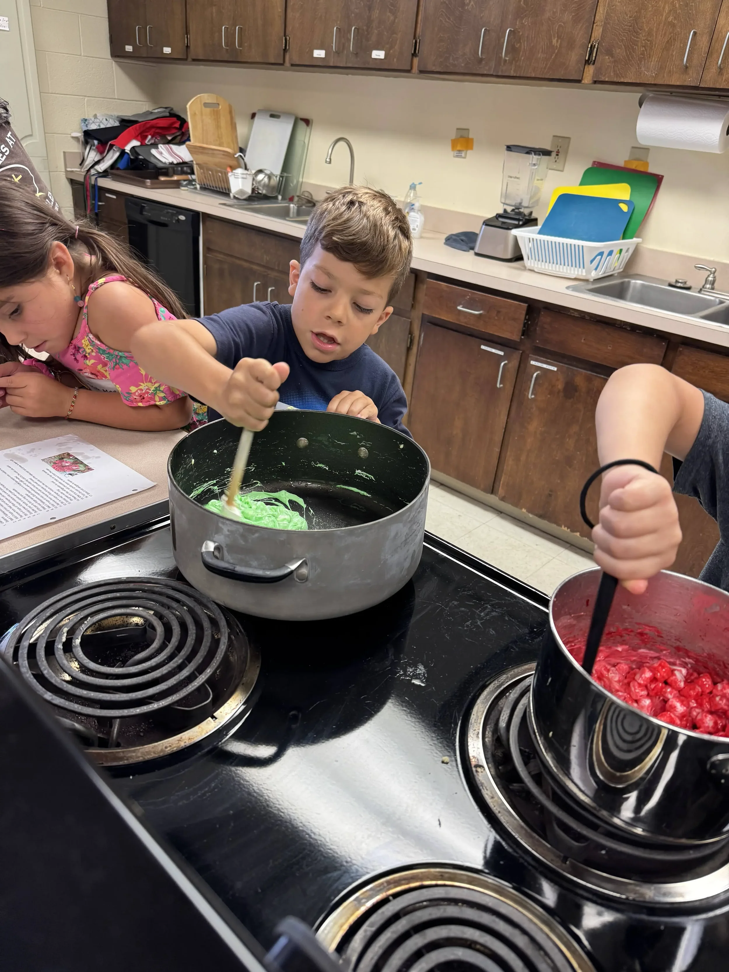  Children stir bright green and red melted ingredients in pots during a hands-on cooking activity. 