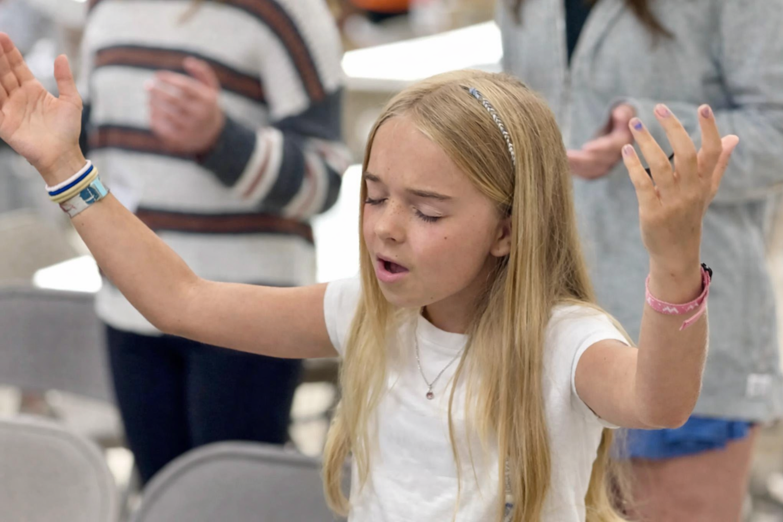 A young girl with eyes closed raises her hands in worship during a group prayer moment.