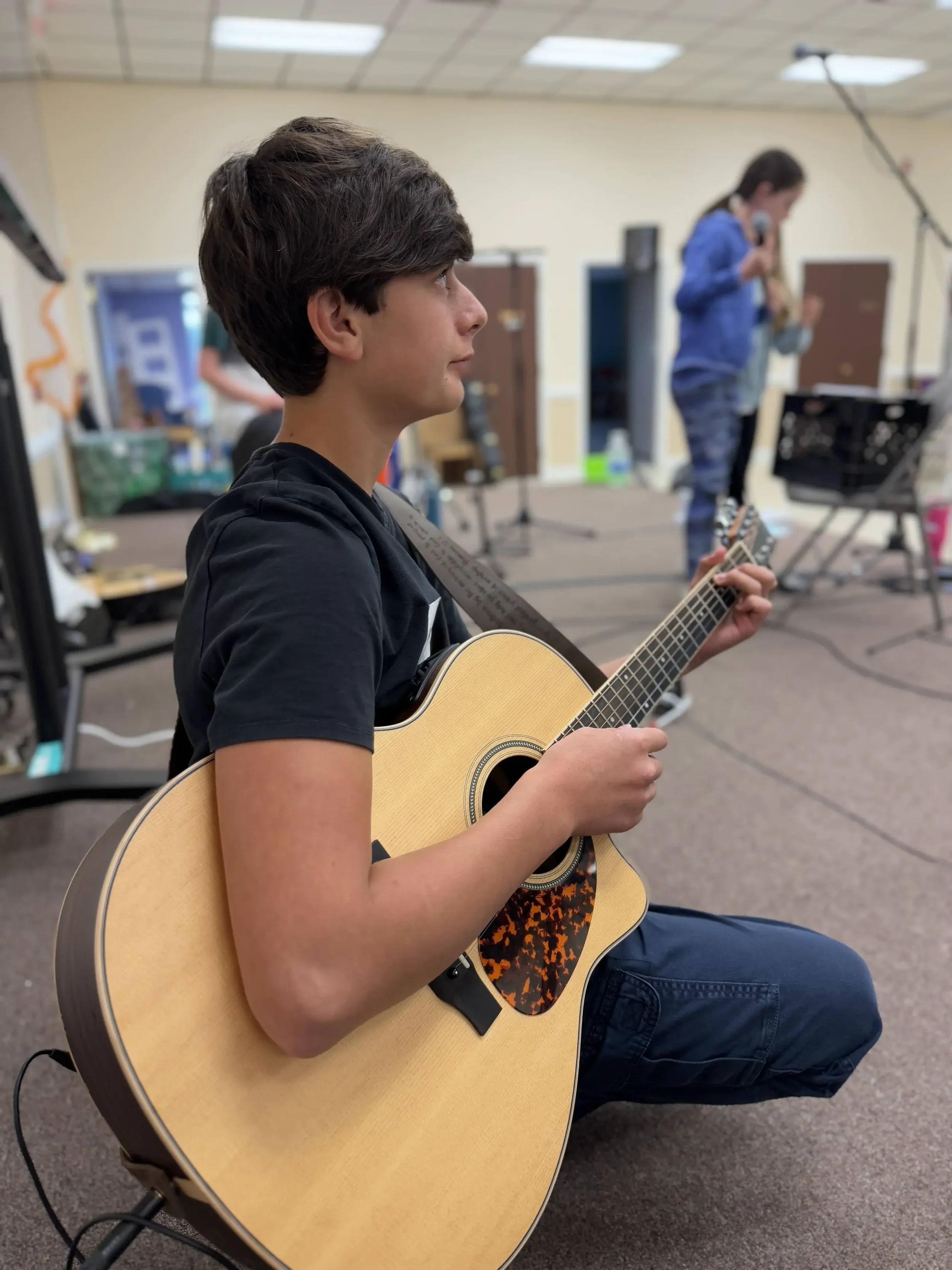 A student sits holding an acoustic guitar, listening attentively during a band rehearsal.