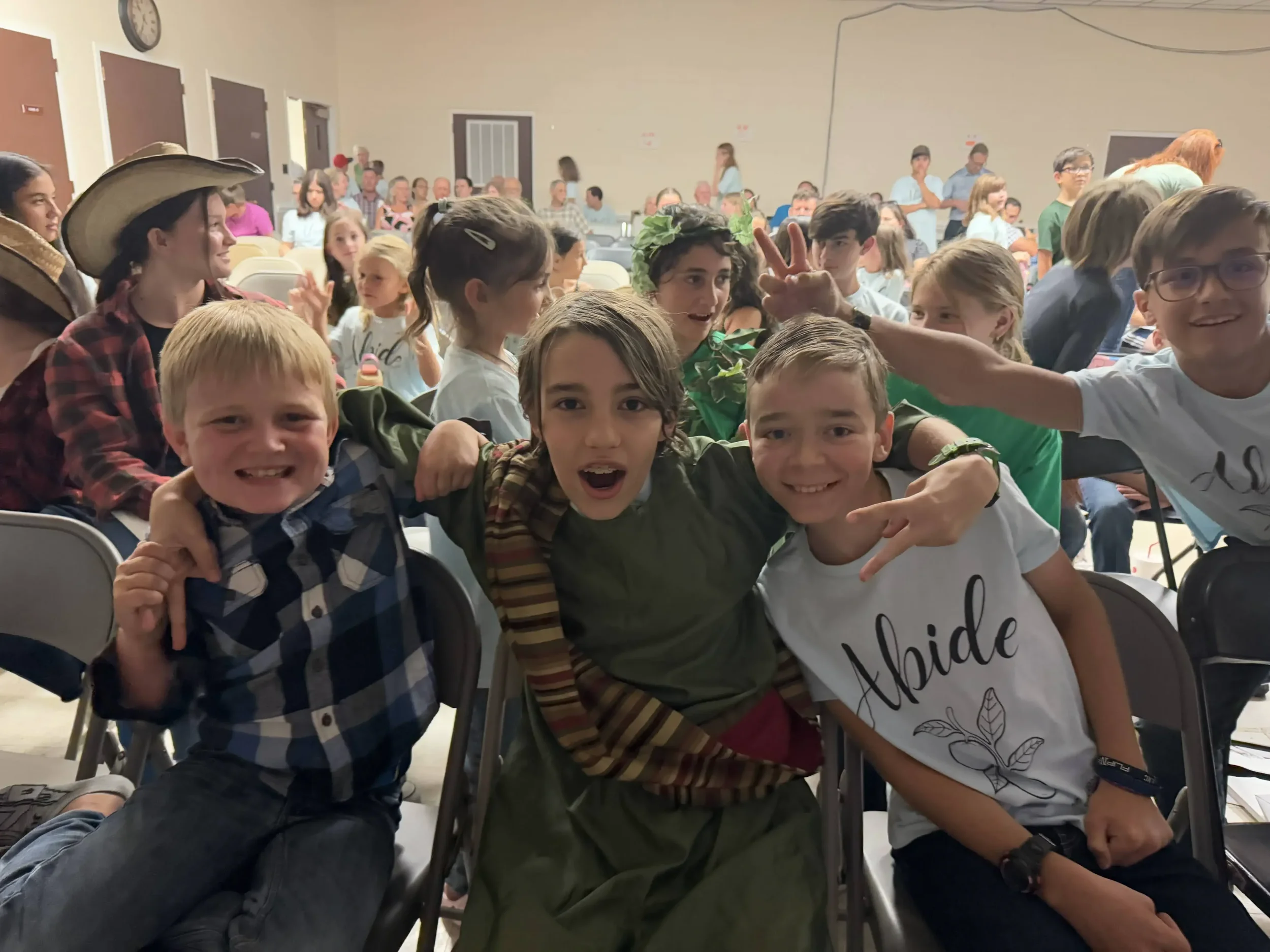 Three smiling boys sit together in an audience, one wearing a costume, posing excitedly.