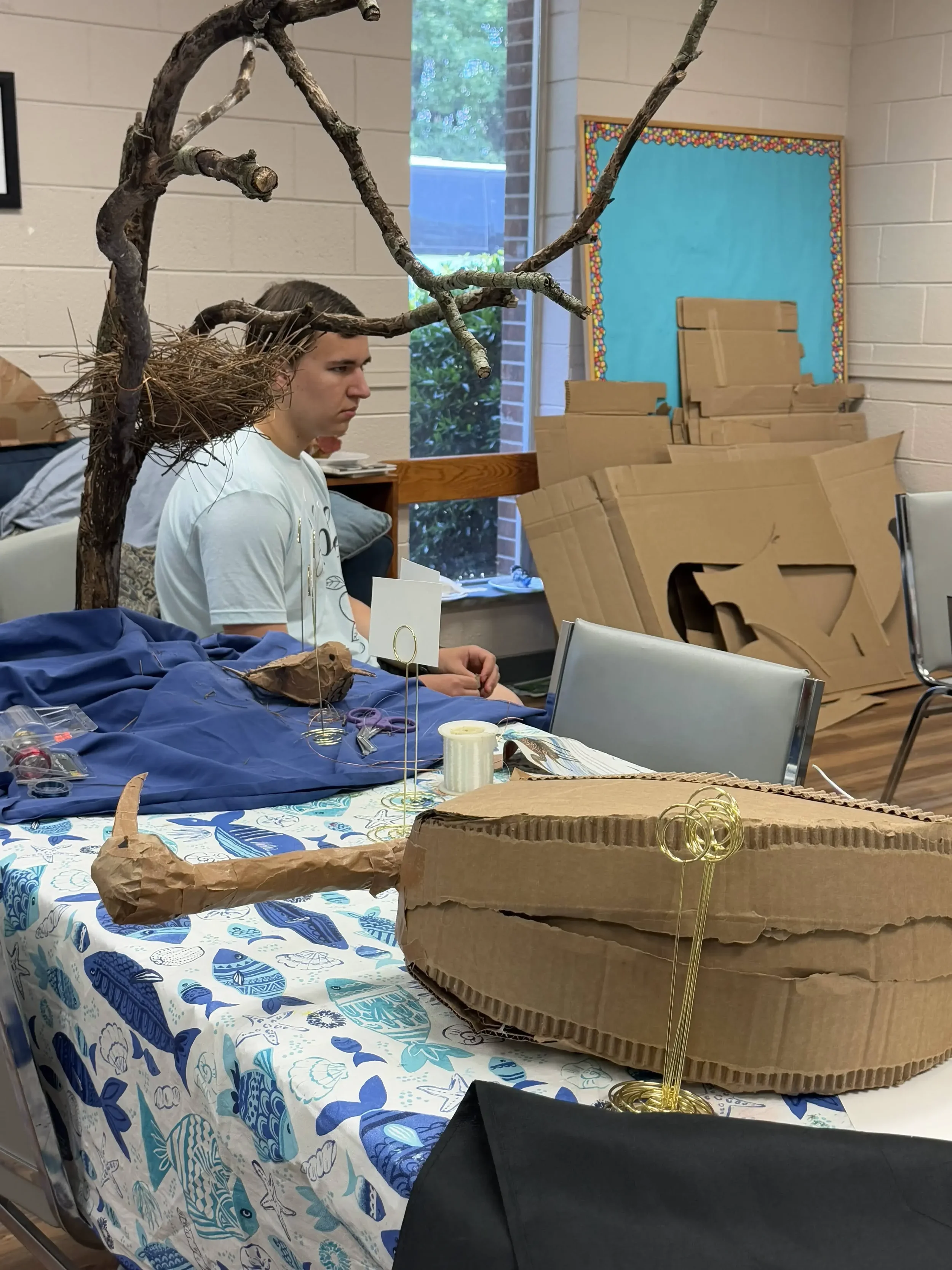A student sits beside a table displaying a nature-themed sculpture made from branches, wire, and cardboard.