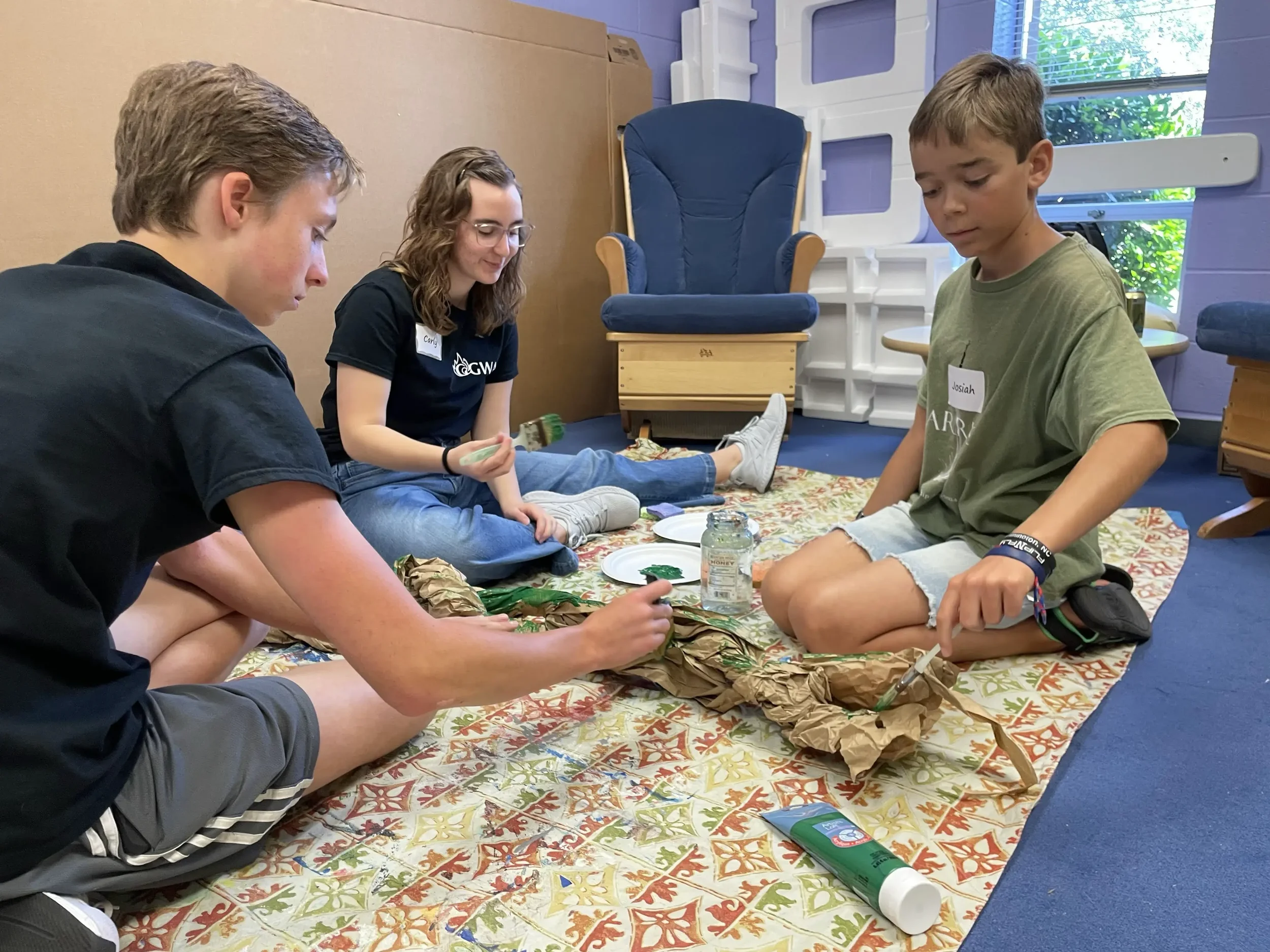 Three students sit on the floor painting and assembling a cardboard prop together during a set design activity.