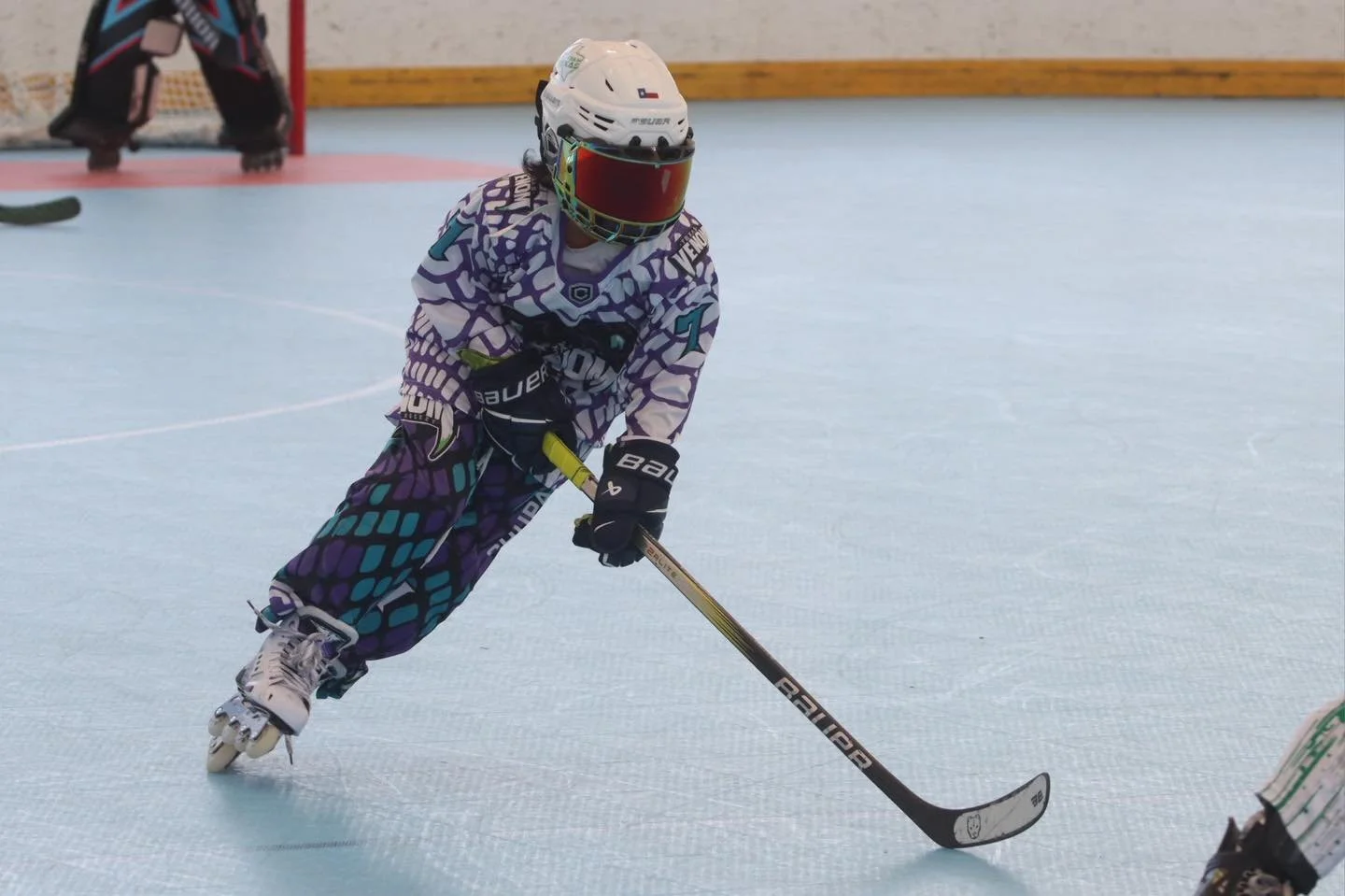 A young person in colorful hockey gear, including a helmet with a visor, sweater, pants, gloves, and skates, playing ice hockey on an indoor rink, handling a hockey stick.