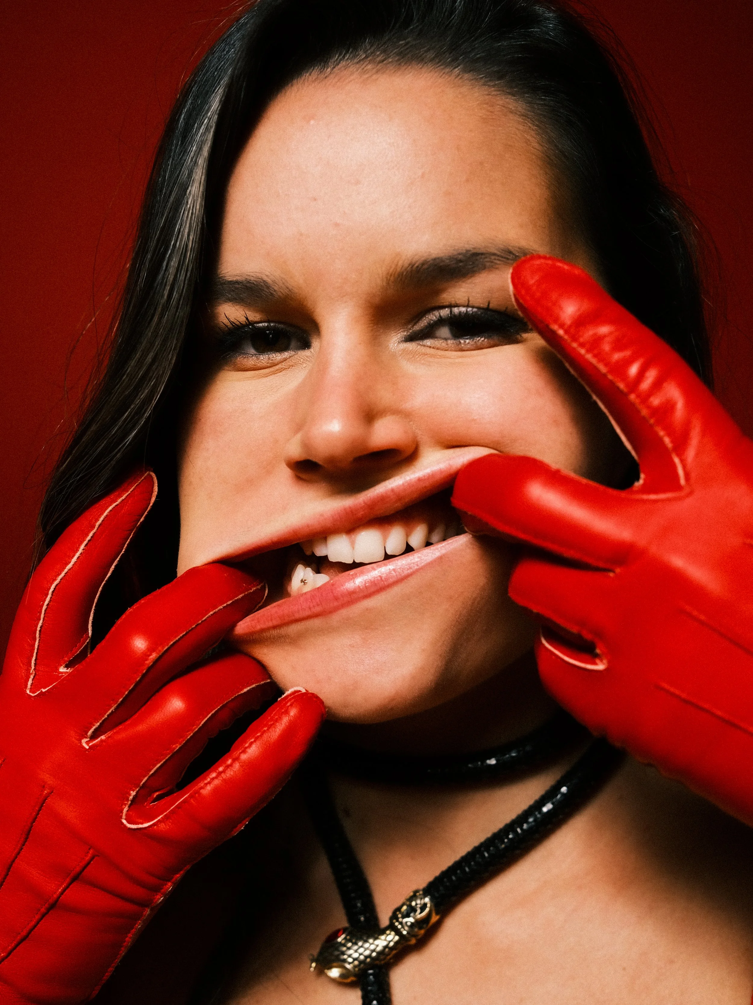 A woman smiling, wearing red leather gloves and a black necklace with a silver clasp, with a red background.