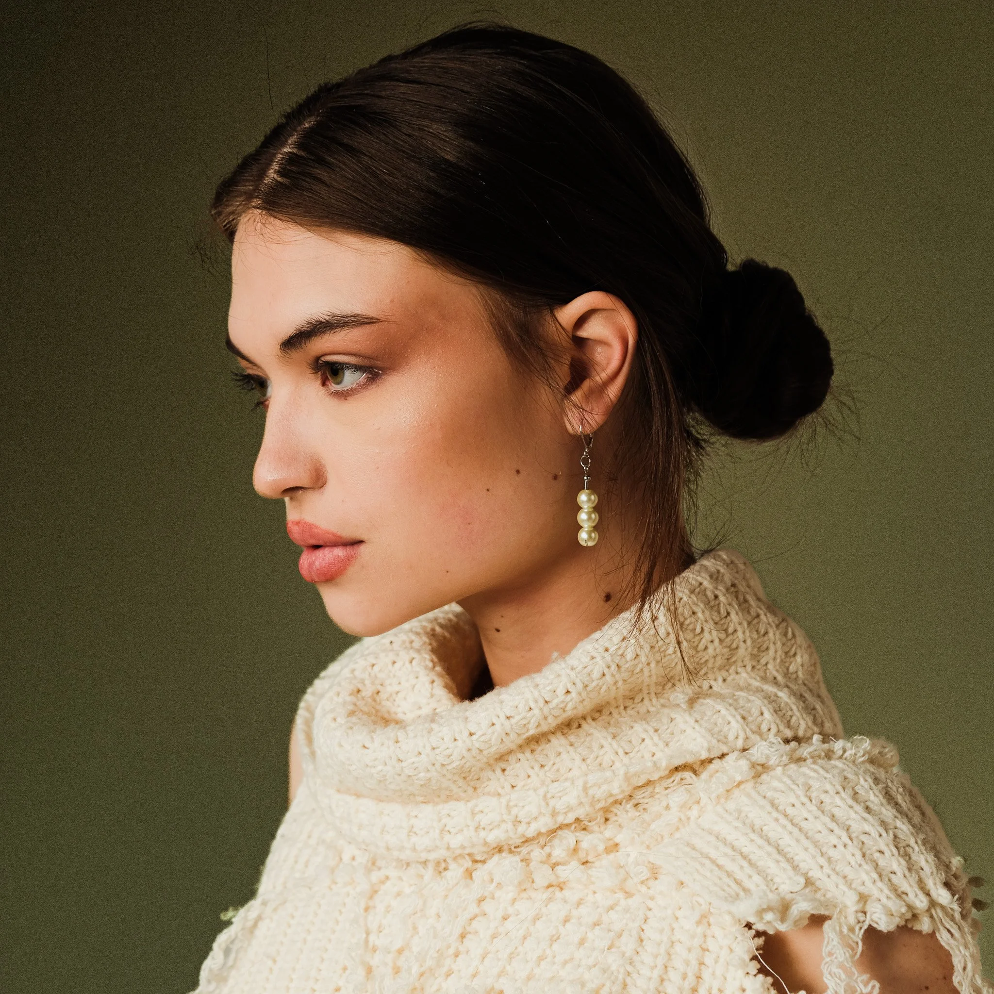 Close-up of a woman's face and side profile showing her eye, eyebrow, and a pearl earring, with she wearing a chunky beige knit sweater, against a plain background.