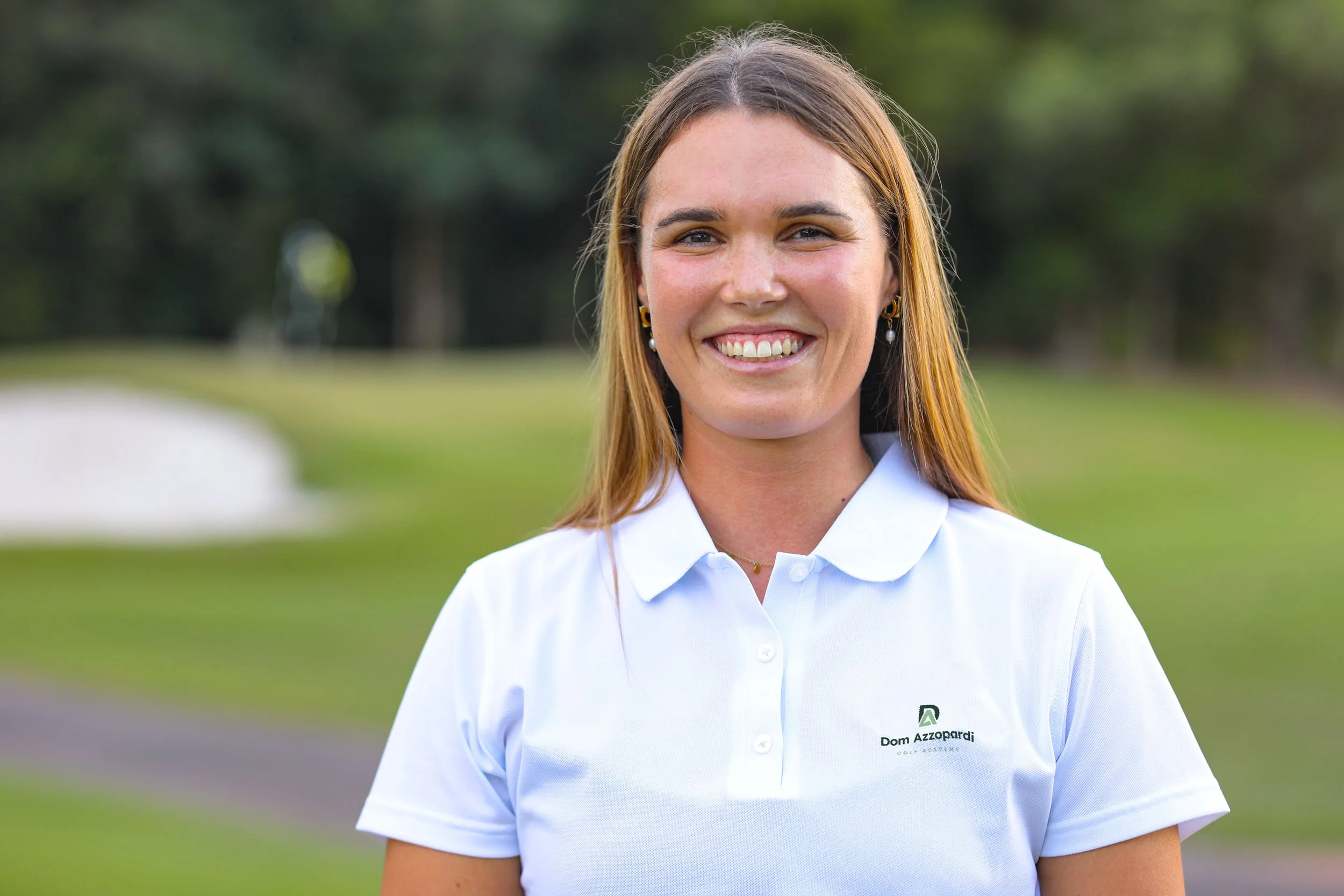 Physiotherapist Laura Neureuter, smiling, with long brown hair, wearing a white polo shirt with a logo that says 'Dom Azzopardi Golf Academy,' standing outdoors on a golf course with green grass and trees in the background.