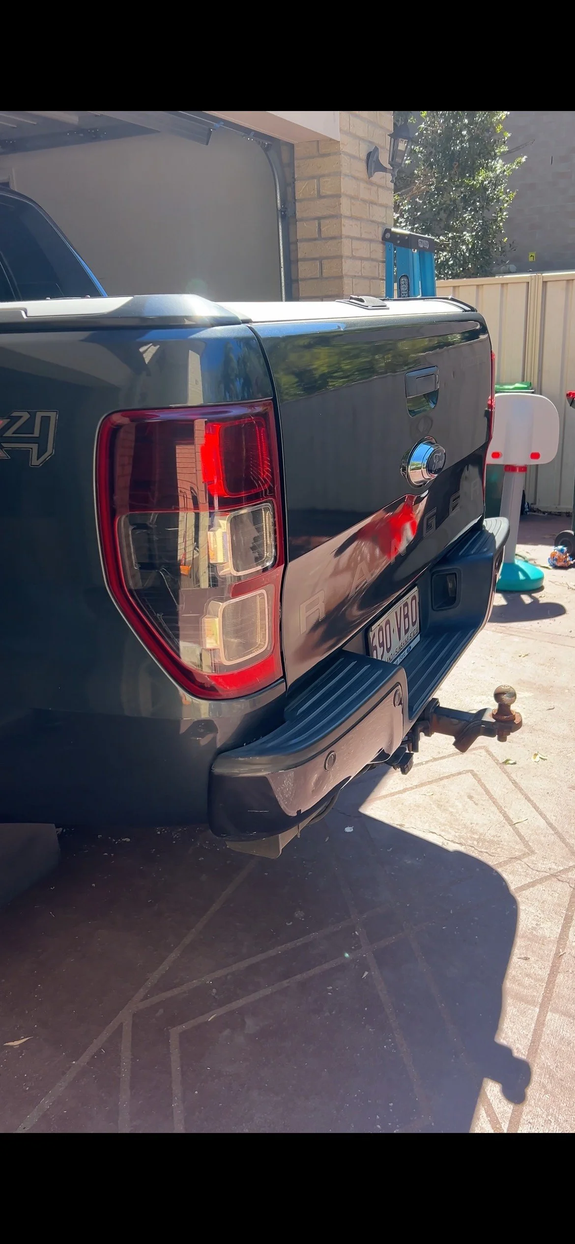 Back of a black pickup truck parked in a driveway, with a spare tire hitch and a license plate visible. The background shows a brick wall, a tree, a white fence, and part of a garage door.