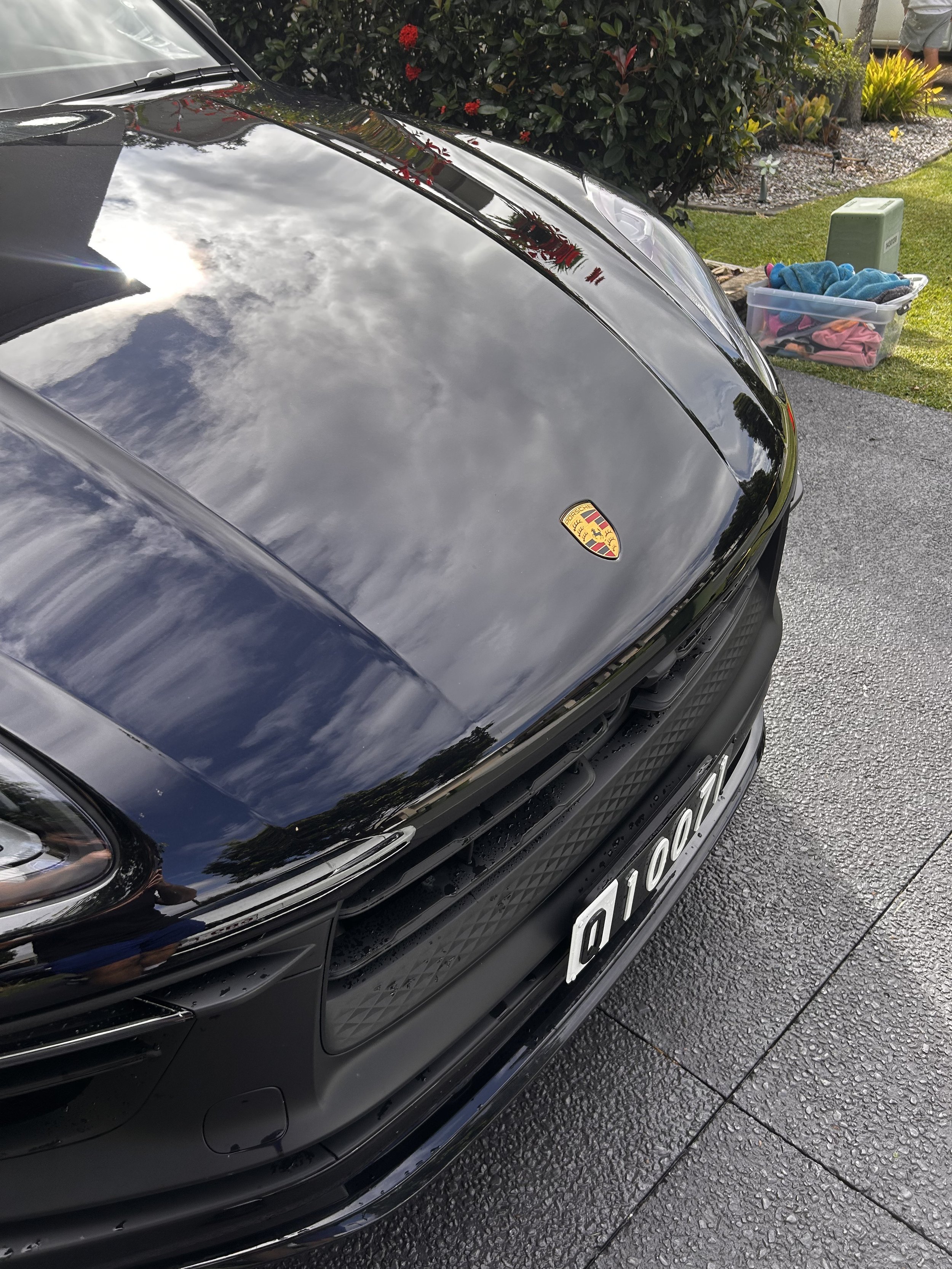 Close-up of the front of a black Porsche car parked on a driveway, with the car's reflective hood showing the cloudy sky and nearby trees.