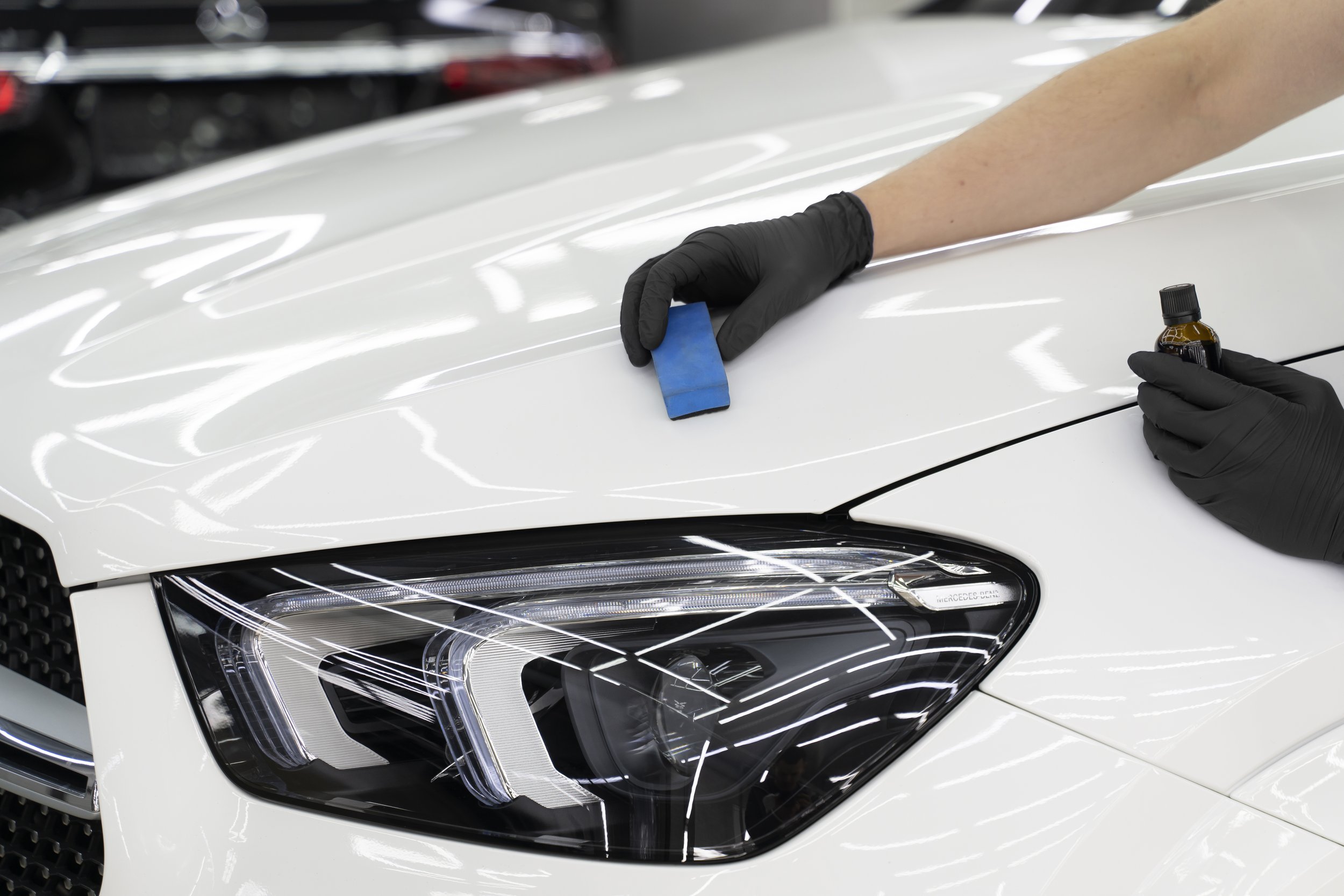 A person wearing black gloves applying a protective coating or polish to a white car's headlight with a blue sponge, holding a small bottle of liquid in one hand, in an auto detailing shop.