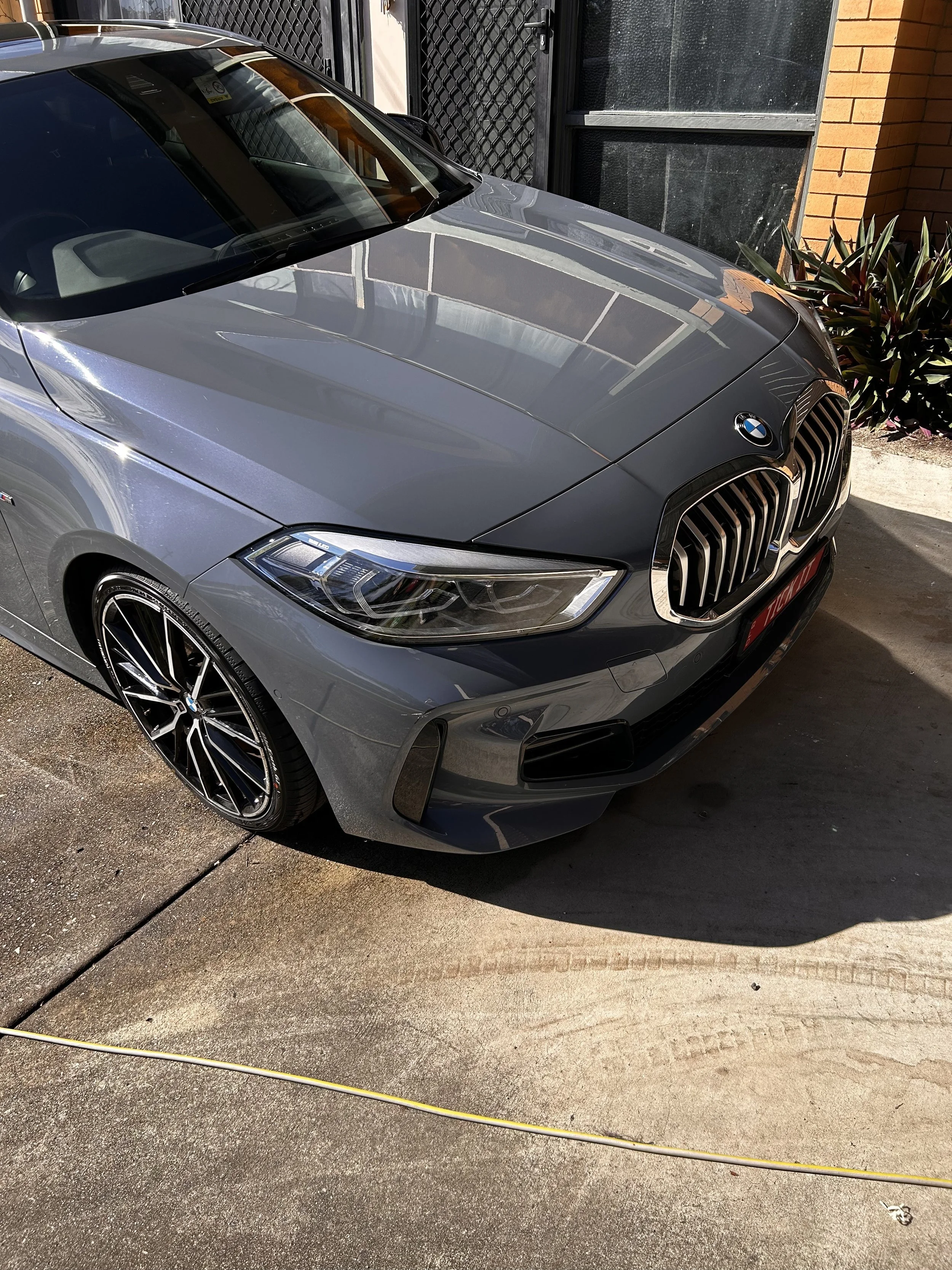 A gray BMW car parked on a concrete driveway, with a brick wall and plants in the background.