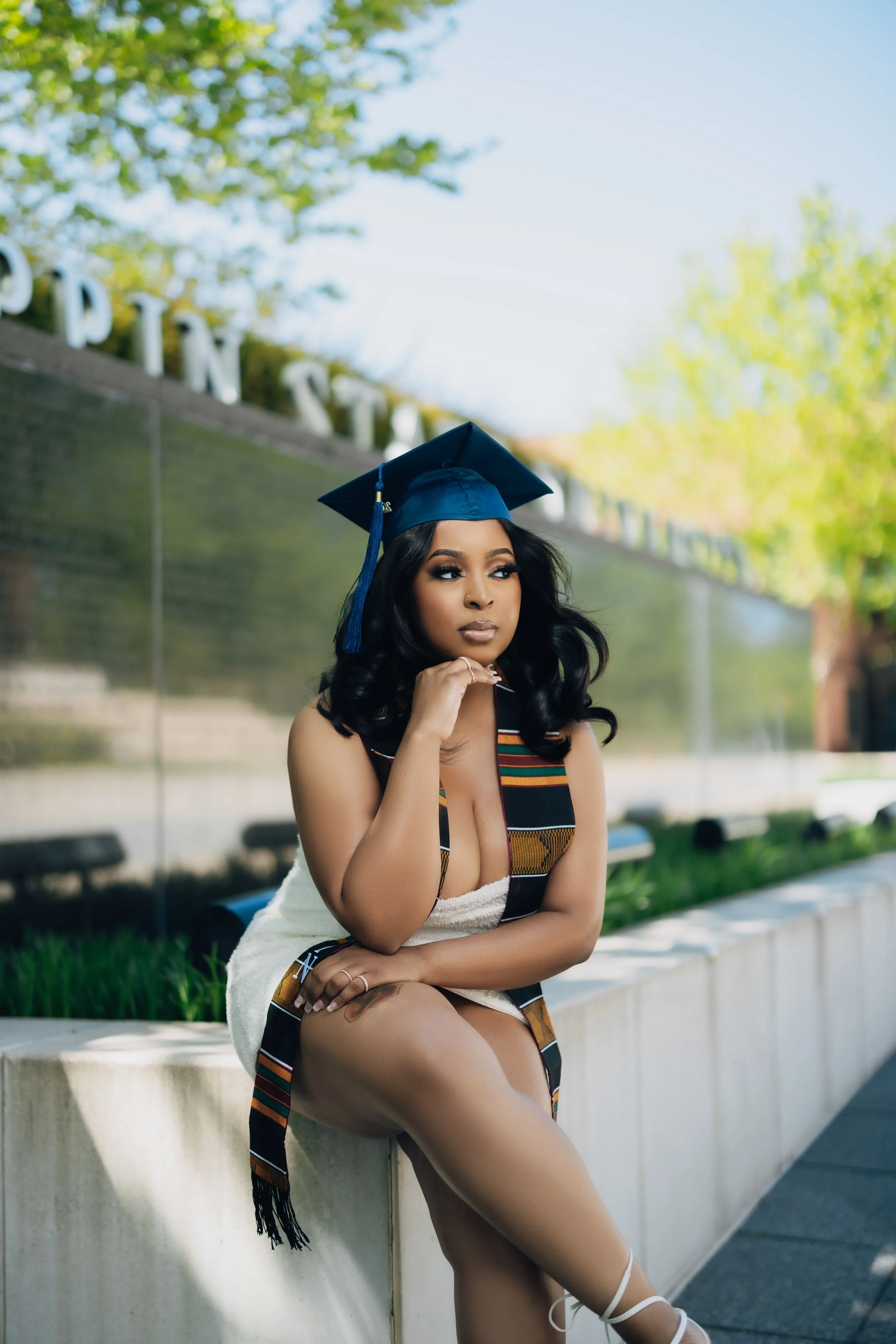 Towson University graduate wearing cap and gown sitting outdoors on a campus ledge with trees in the background