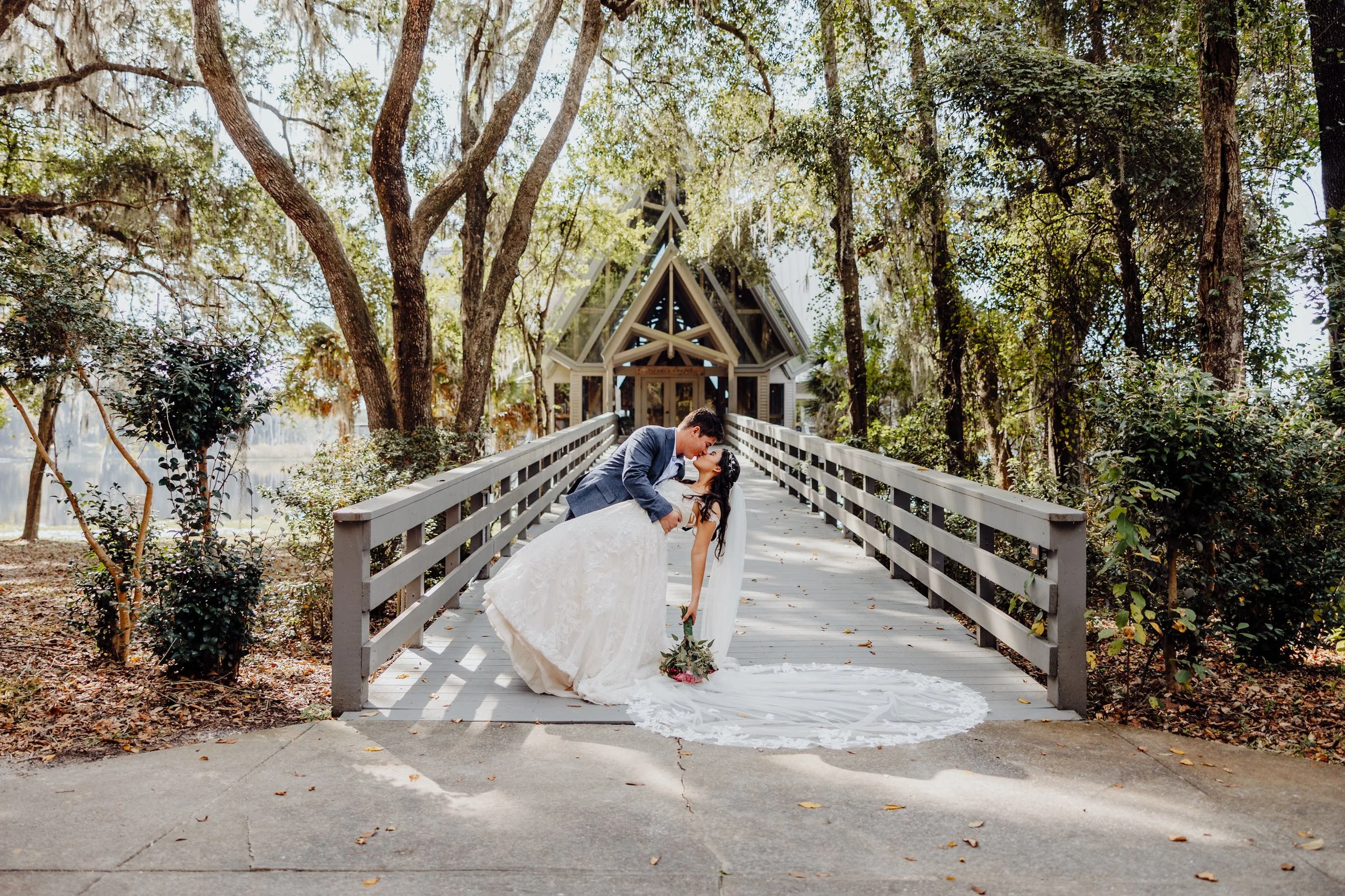 Bride and groom kissing on a path, surrounded by trees and a chapel in the background.