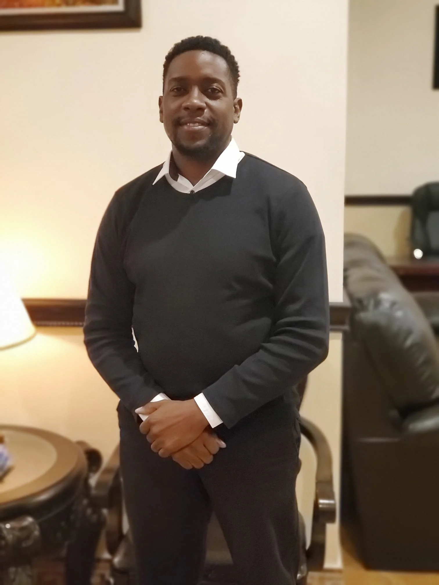 A man standing indoors wearing a white collared shirt and black sweater, with a beige wall and furniture in the background.