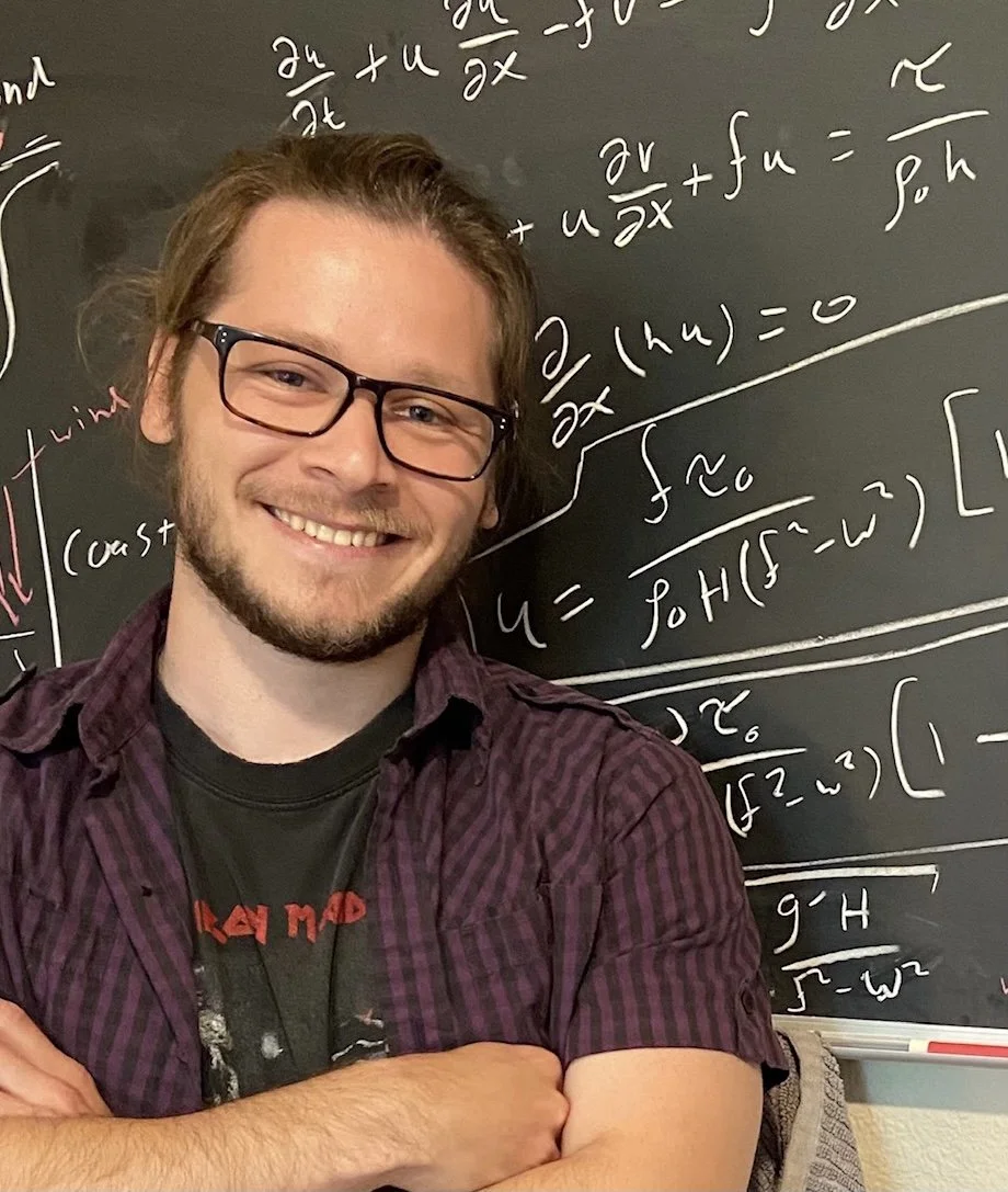A man with glasses and a beard smiling, standing in front of a chalkboard filled with mathematical equations.
