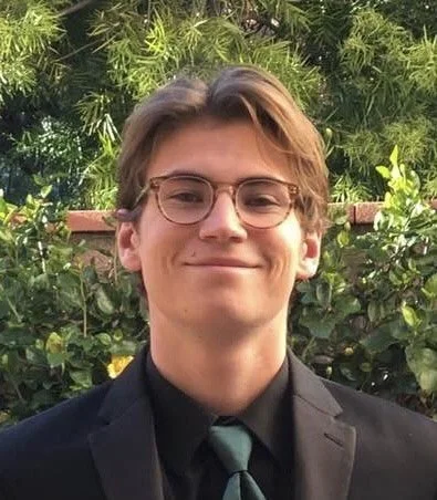 A young man with light brown hair and glasses, wearing a suit and tie, standing outdoors with green foliage in the background.