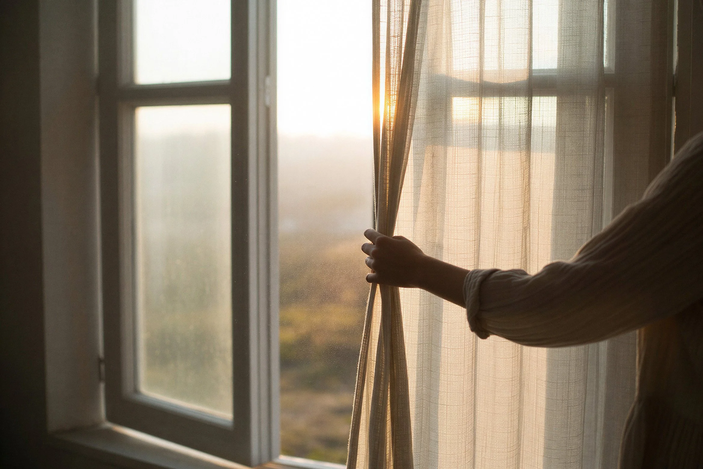 woman's arm opening curtain on open window to reveal sunlight and view of countryside