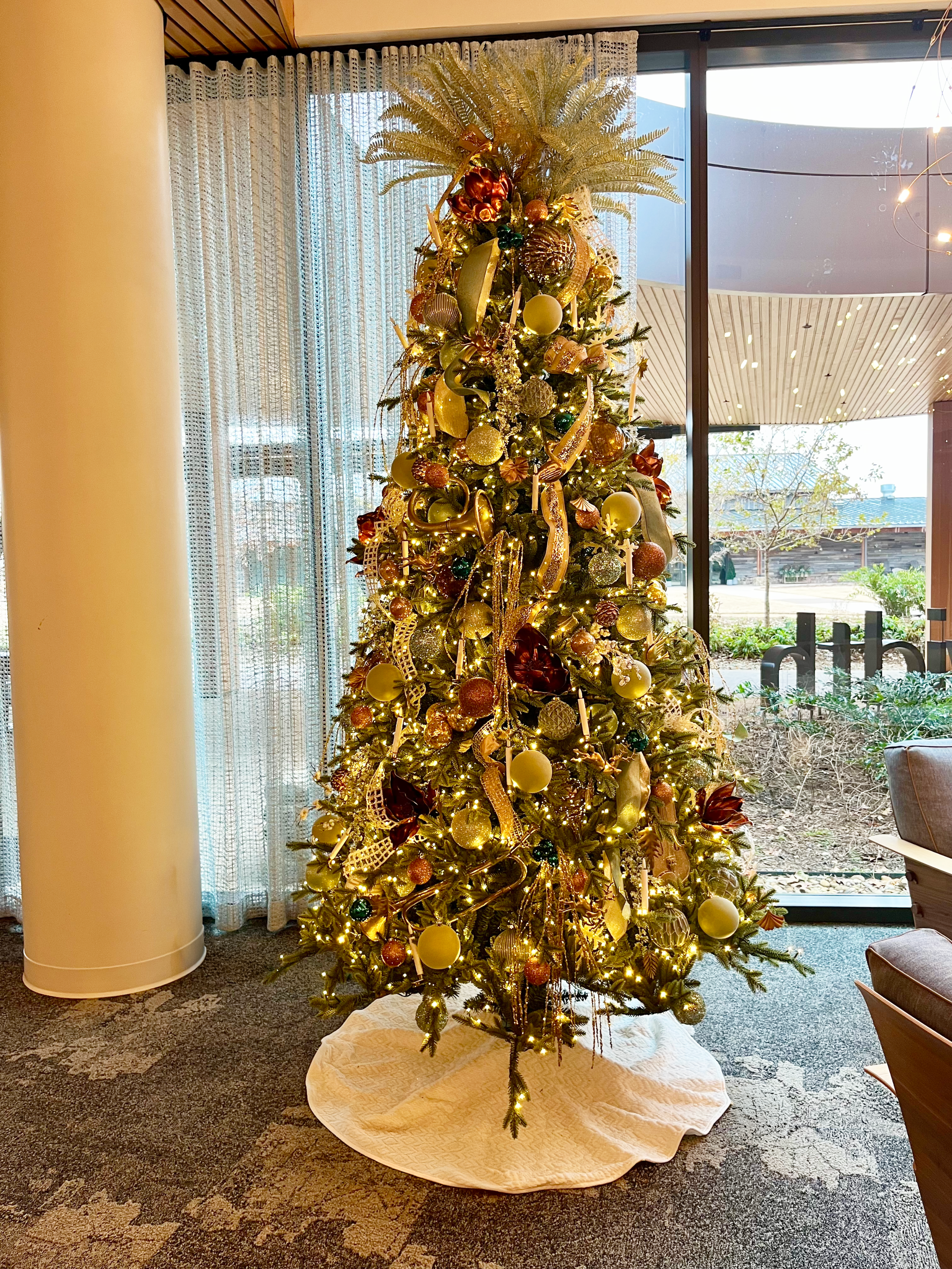 A tall Christmas tree decorated with gold and bronze ornaments, ribbons, and lights, placed on a white tree skirt inside a room with large windows and curtains.