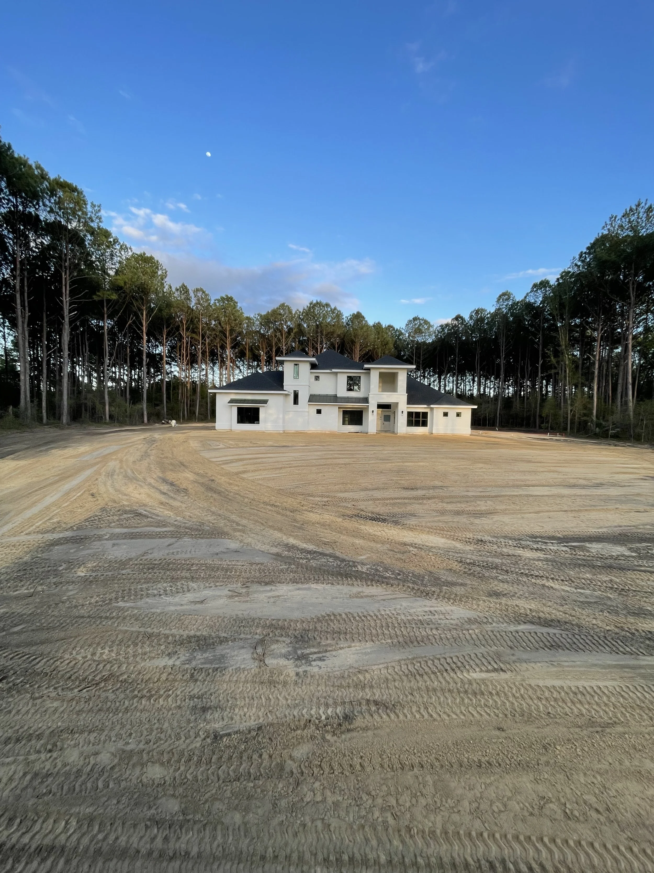 A large, unfinished house on a plot of land surrounded by tall trees under a blue sky.