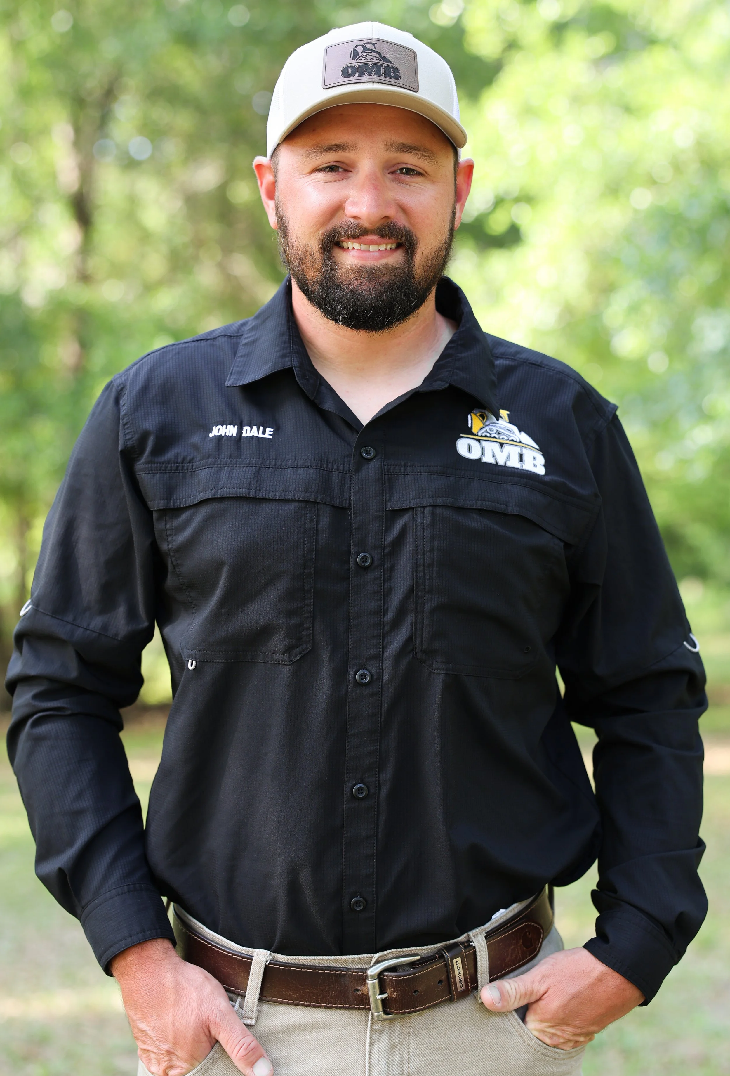 Man in a black collared shirt and tan baseball cap standing outdoors with trees in the background.