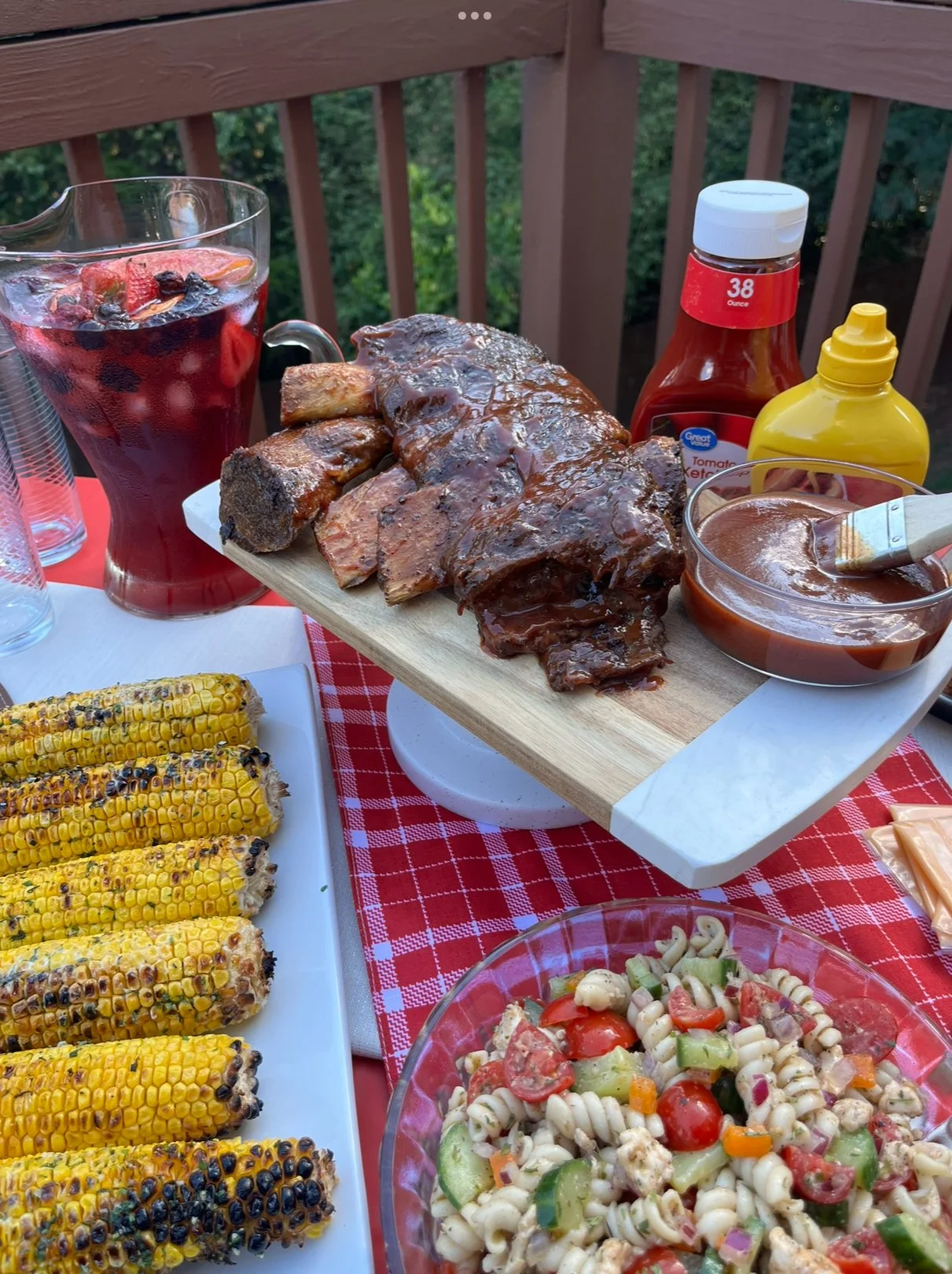 Barbecue meal with grilled corn on the cob, sliced brisket ribs with barbecue sauce on a serving board, pasta salad with vegetables, and a glass of red berry drink, set on a red and white checkered tablecloth on a wooden deck.