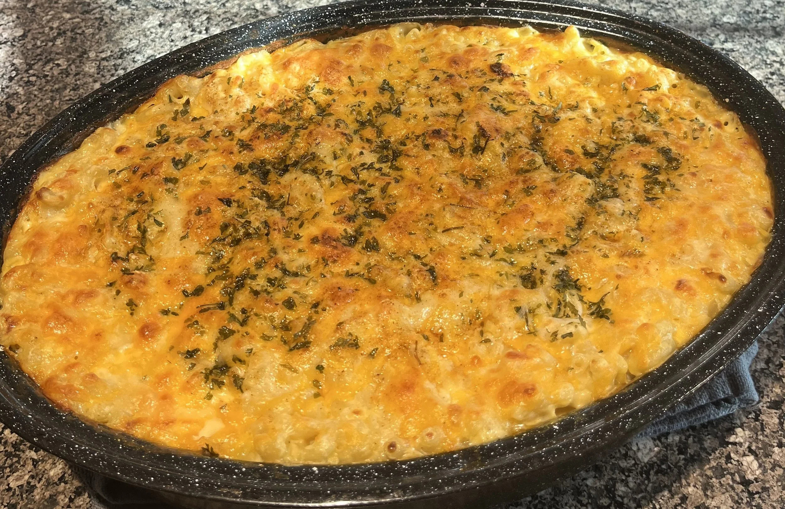 A baked dish in a black oval casserole dish, topped with melted cheese and garnished with chopped herbs, on a speckled countertop.