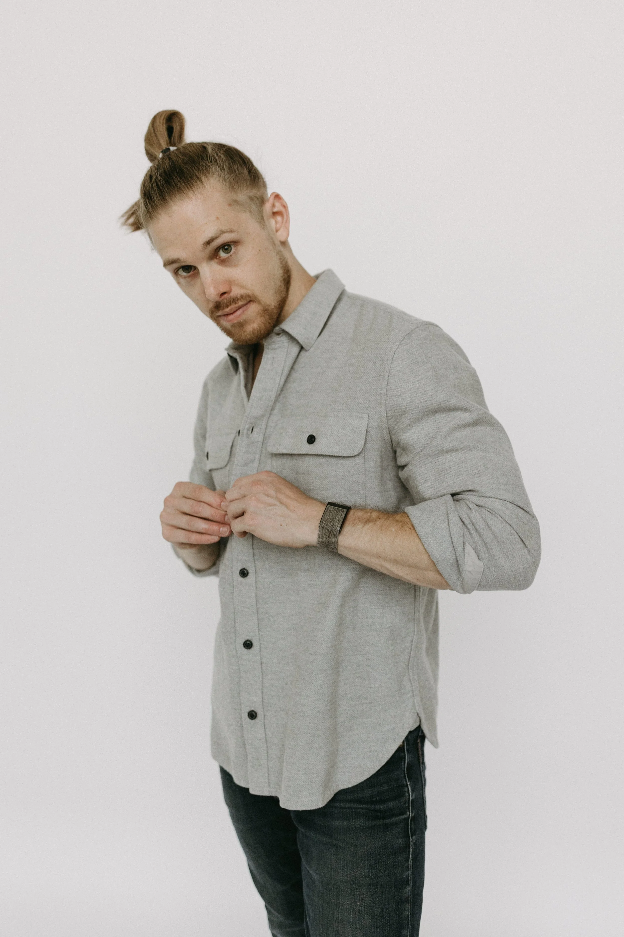 A young man with long hair tied in a bun, wearing a light gray button-up shirt and dark jeans, adjusting his shirt while looking into the camera against a plain white background.