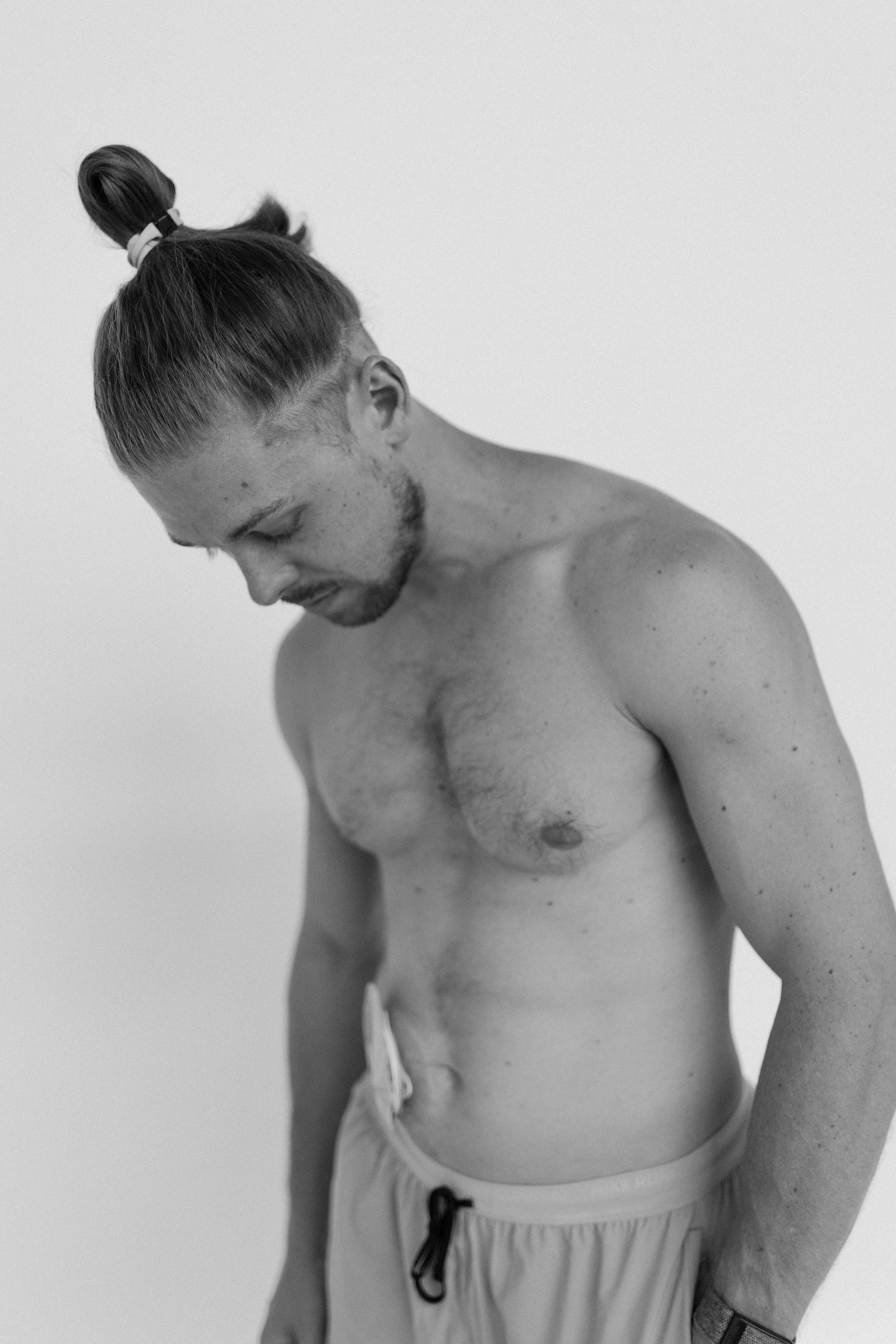 Black-and-white photo of a shirtless young man with long hair tied in a bun looking down, standing against a plain white background.