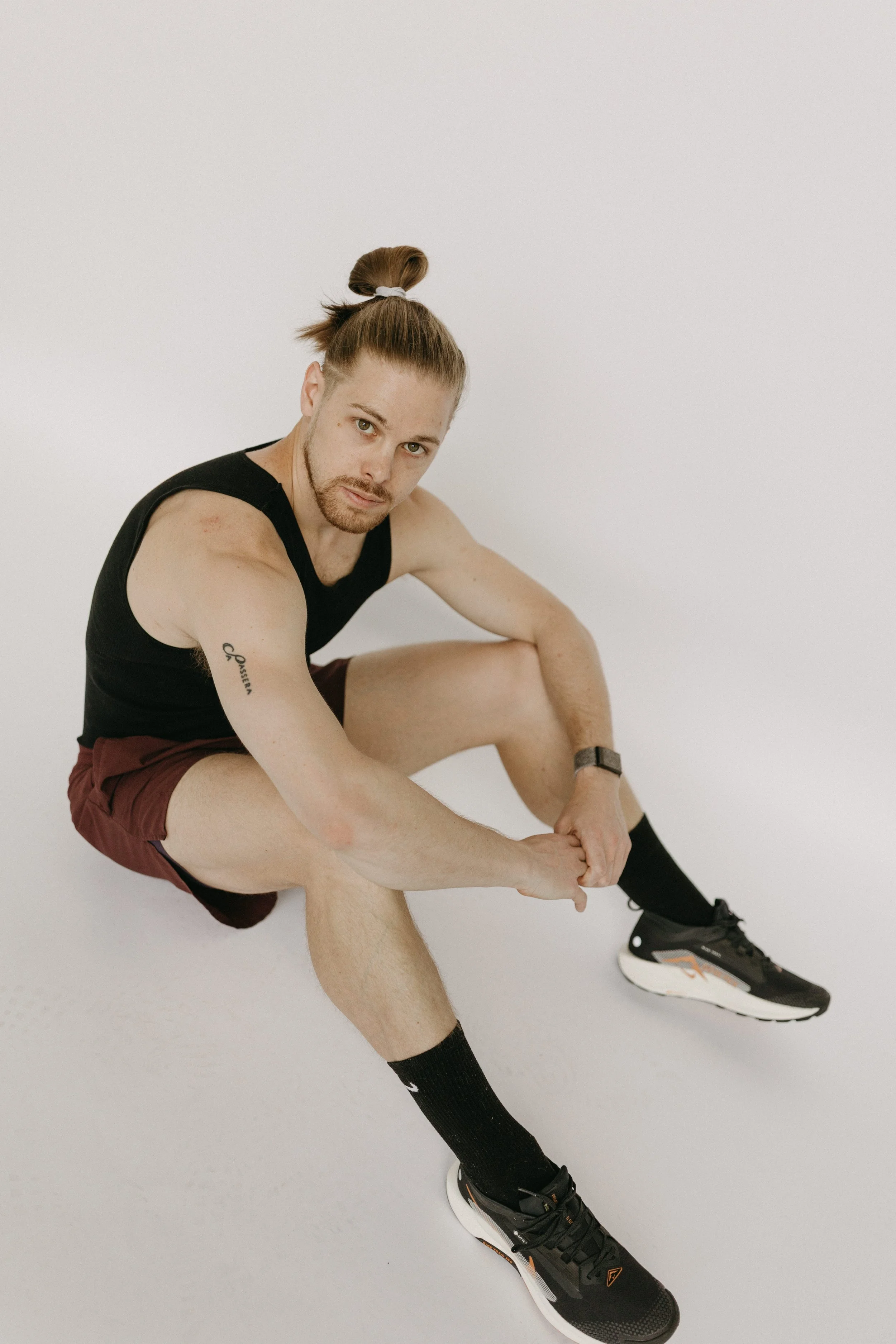 A man sitting on the floor, adjusting his black athletic shoe, wearing a black sleeveless shirt, maroon shorts, black socks, and a watch, with a ponytail hairstyle, against a plain white background.