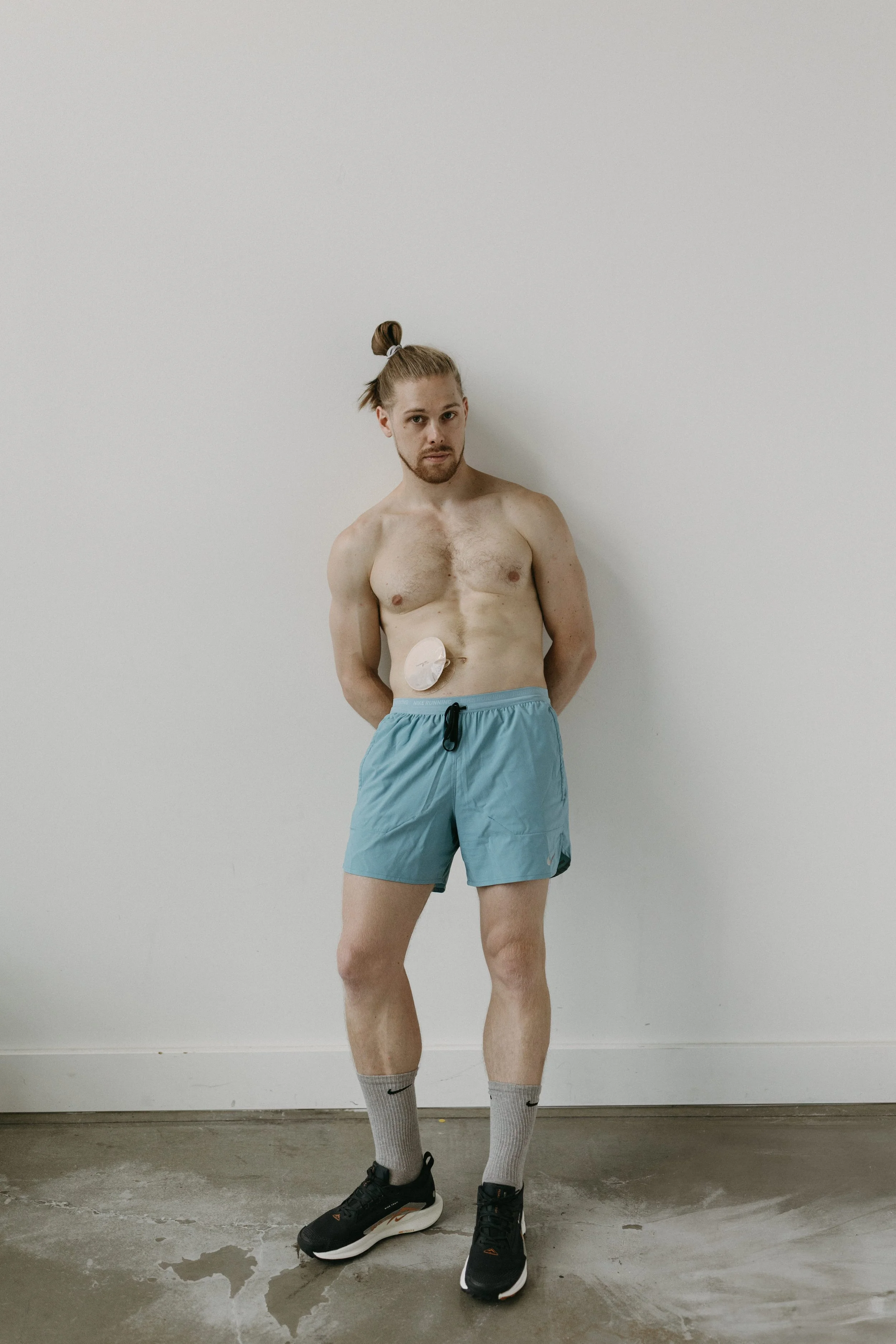 A shirtless man with long hair tied back, standing against a plain white wall, wearing light blue shorts, gray Nike socks, and black athletic shoes.