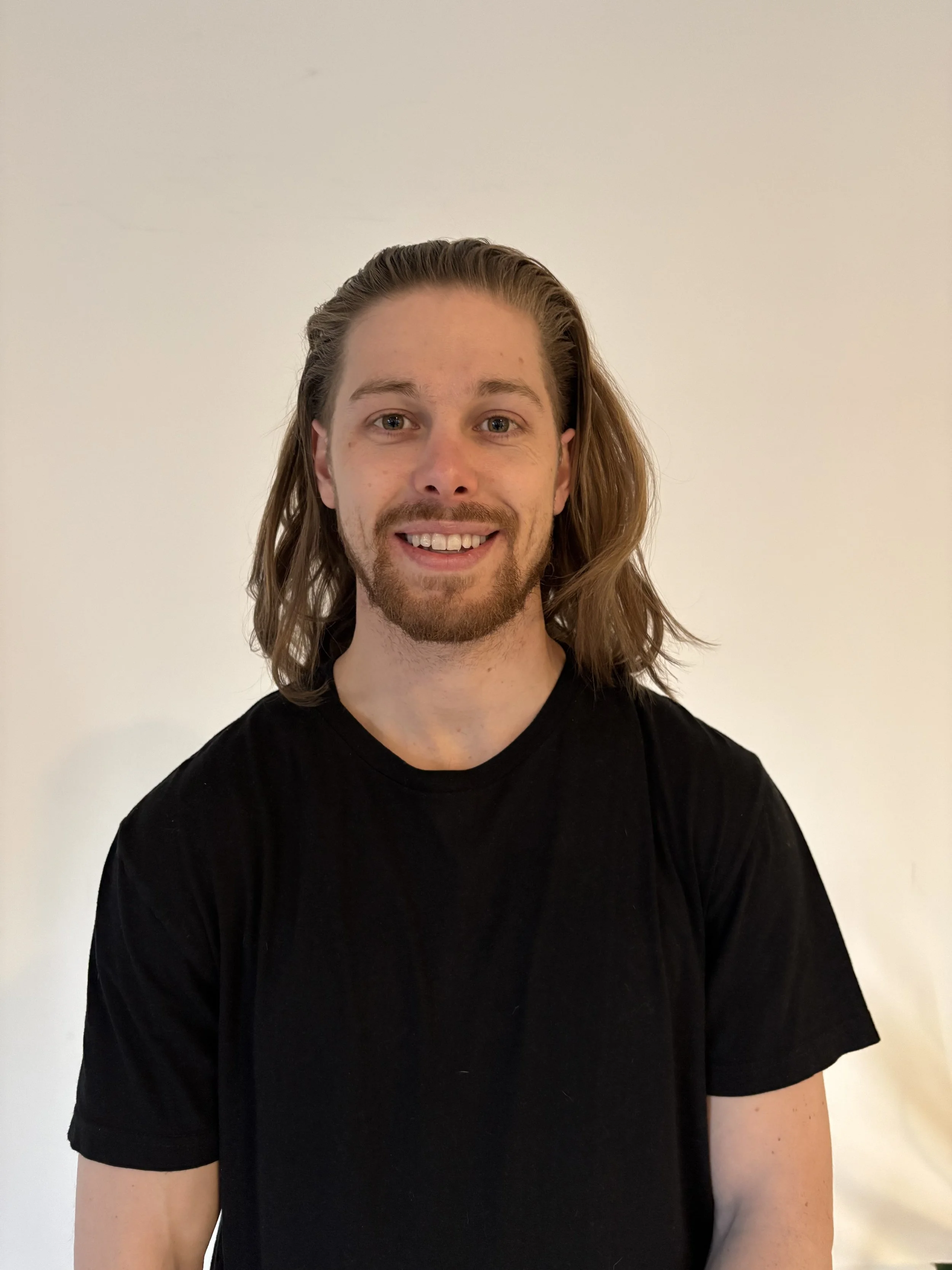 Young man with long brown hair and a beard, smiling, wearing a black t-shirt against a plain light-colored background.