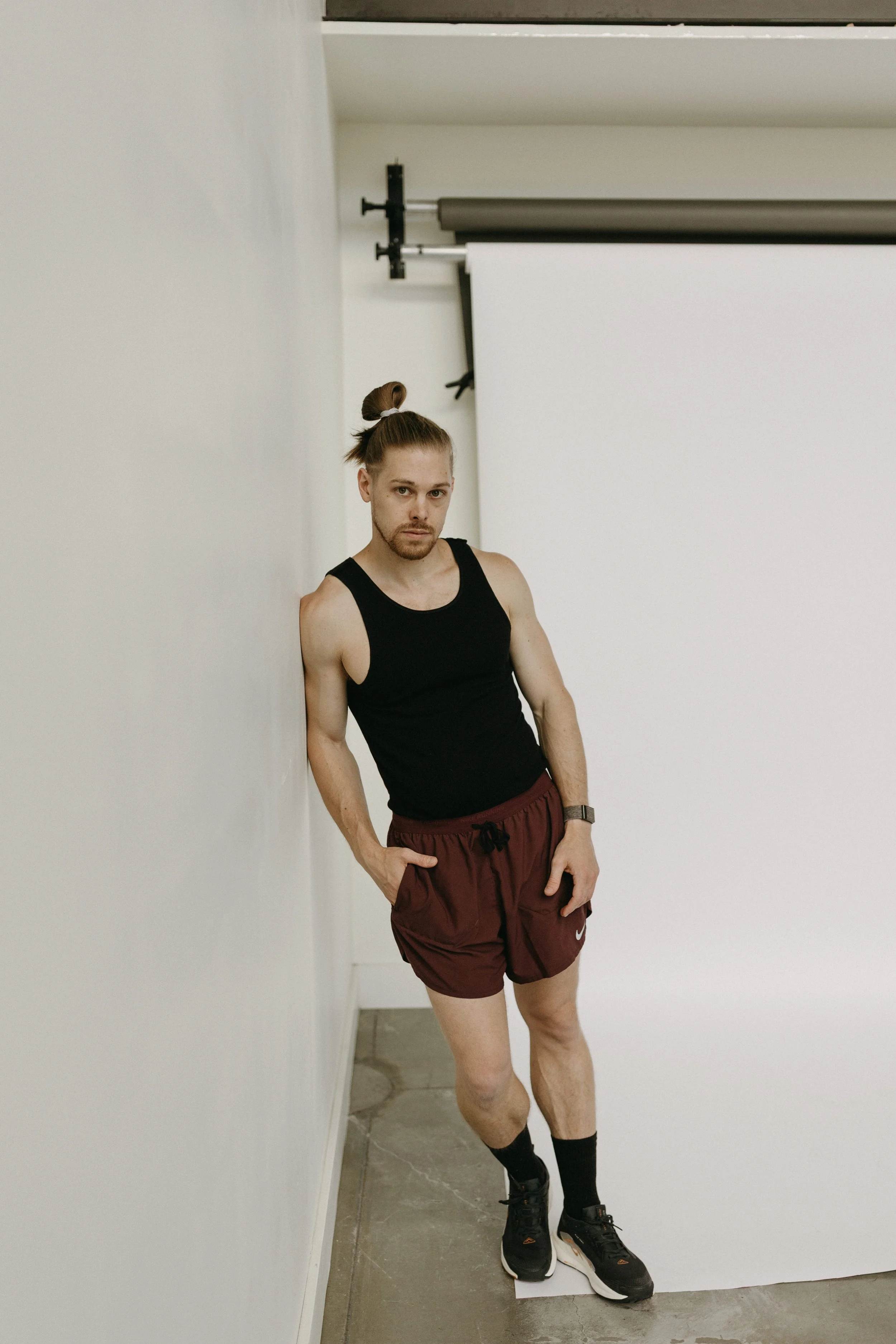 A man with a man bun, black tank top, maroon Nike shorts, and black sneakers leaning against a white wall in a photo studio, with a plain white backdrop and studio equipment visible.
