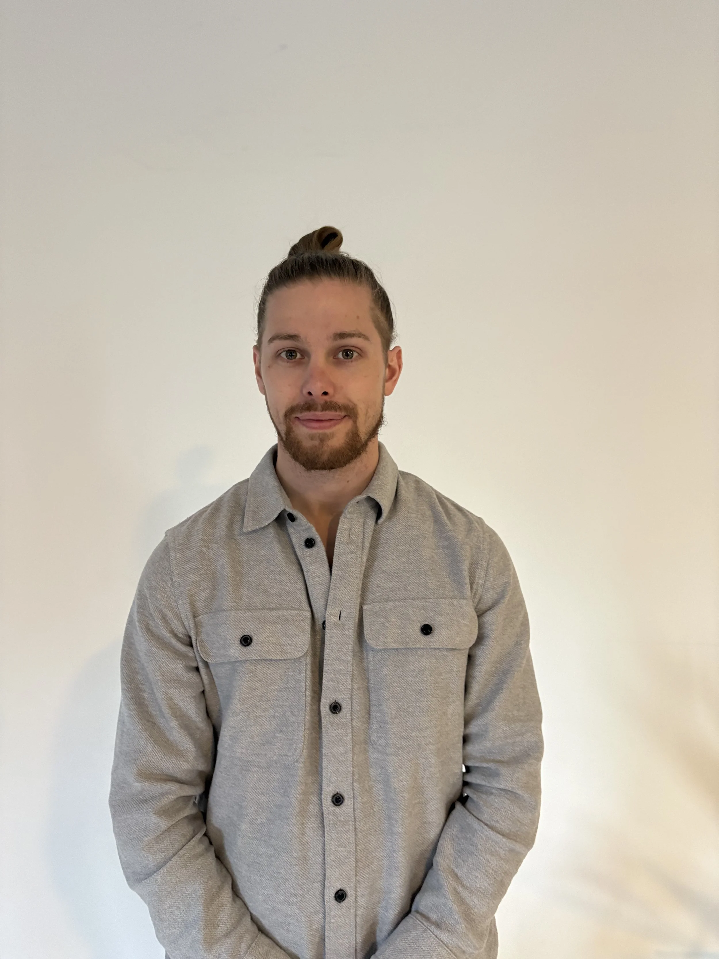 A young man with a beard and mustache, wearing a gray button-up shirt, stands against a plain white wall. His hair is tied up in a bun, and he has a slight smile.