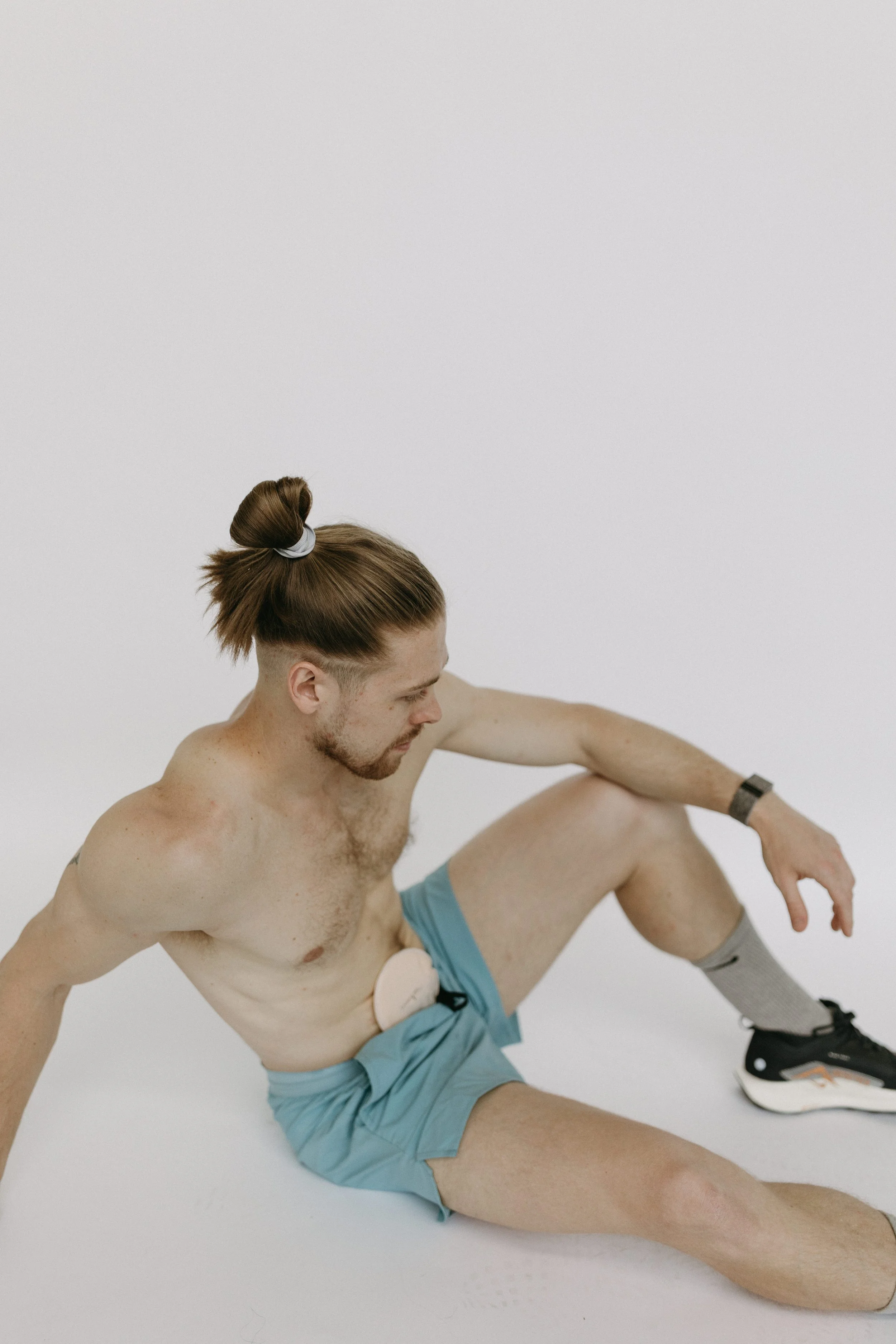 A shirtless man with long hair tied in a bun, sitting on the ground in a white room, wearing blue shorts, gray socks, and black sneakers.