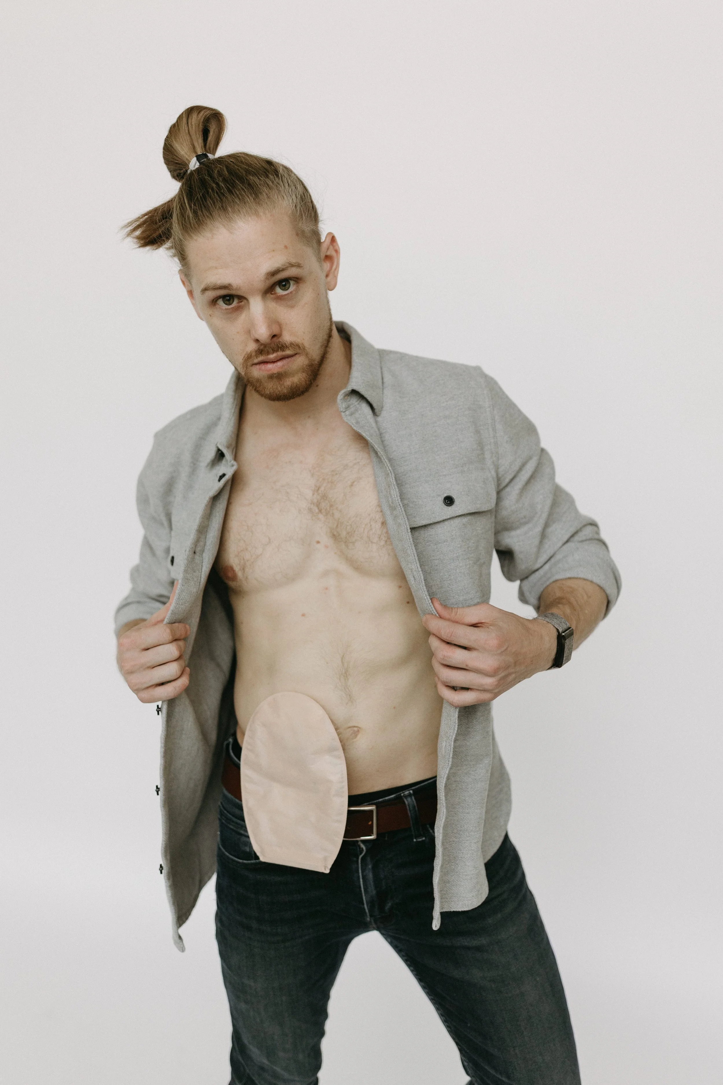 A man with long hair in a ponytail, wearing a gray shirt unbuttoned, showing his bare chest and torso with an ileostomy bag. He is holding open his shirt with both hands and looking directly at the camera.