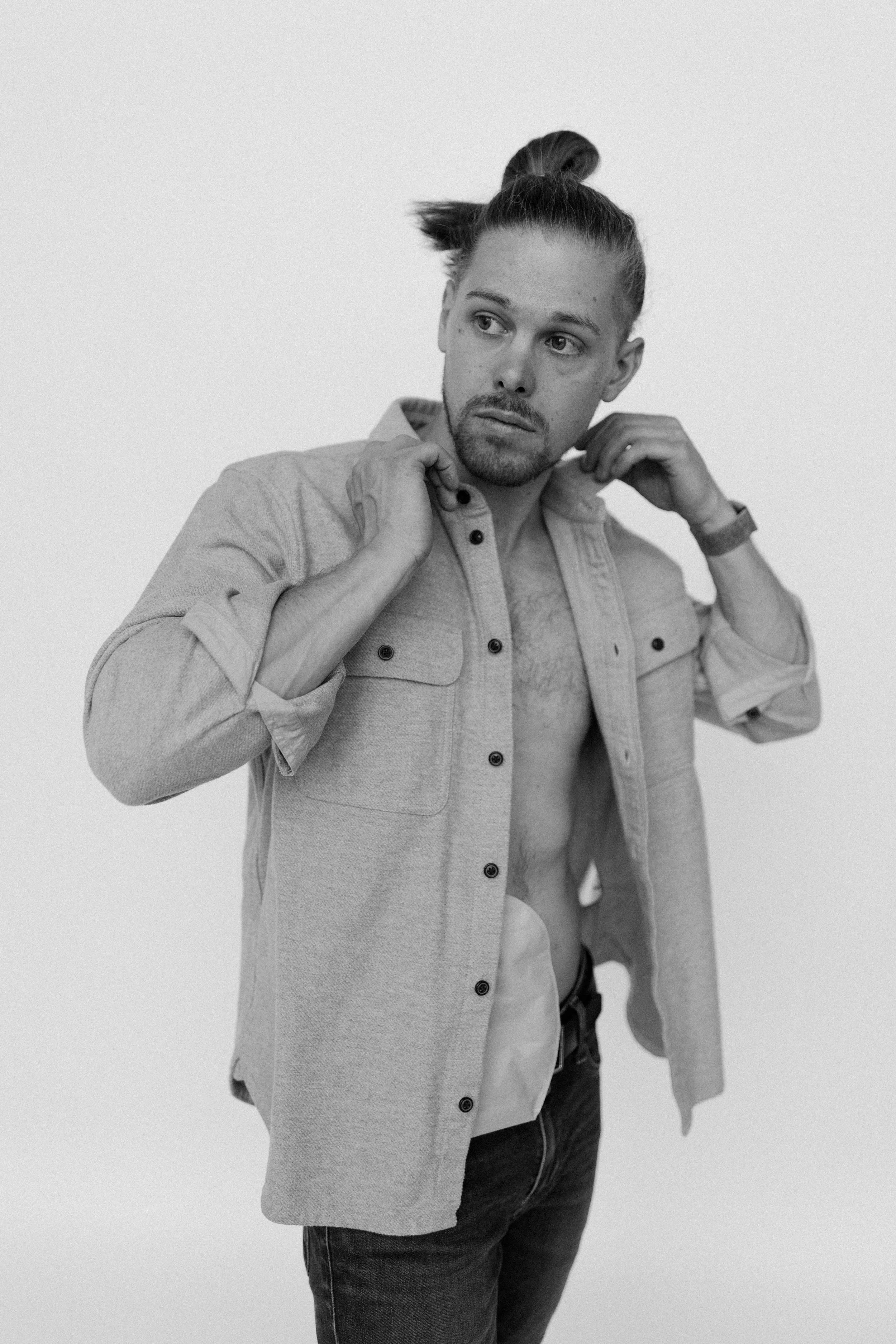 A man with a beard and long hair in a bun, adjusting his collar while wearing an open jacket exposing his chest, against a plain background.