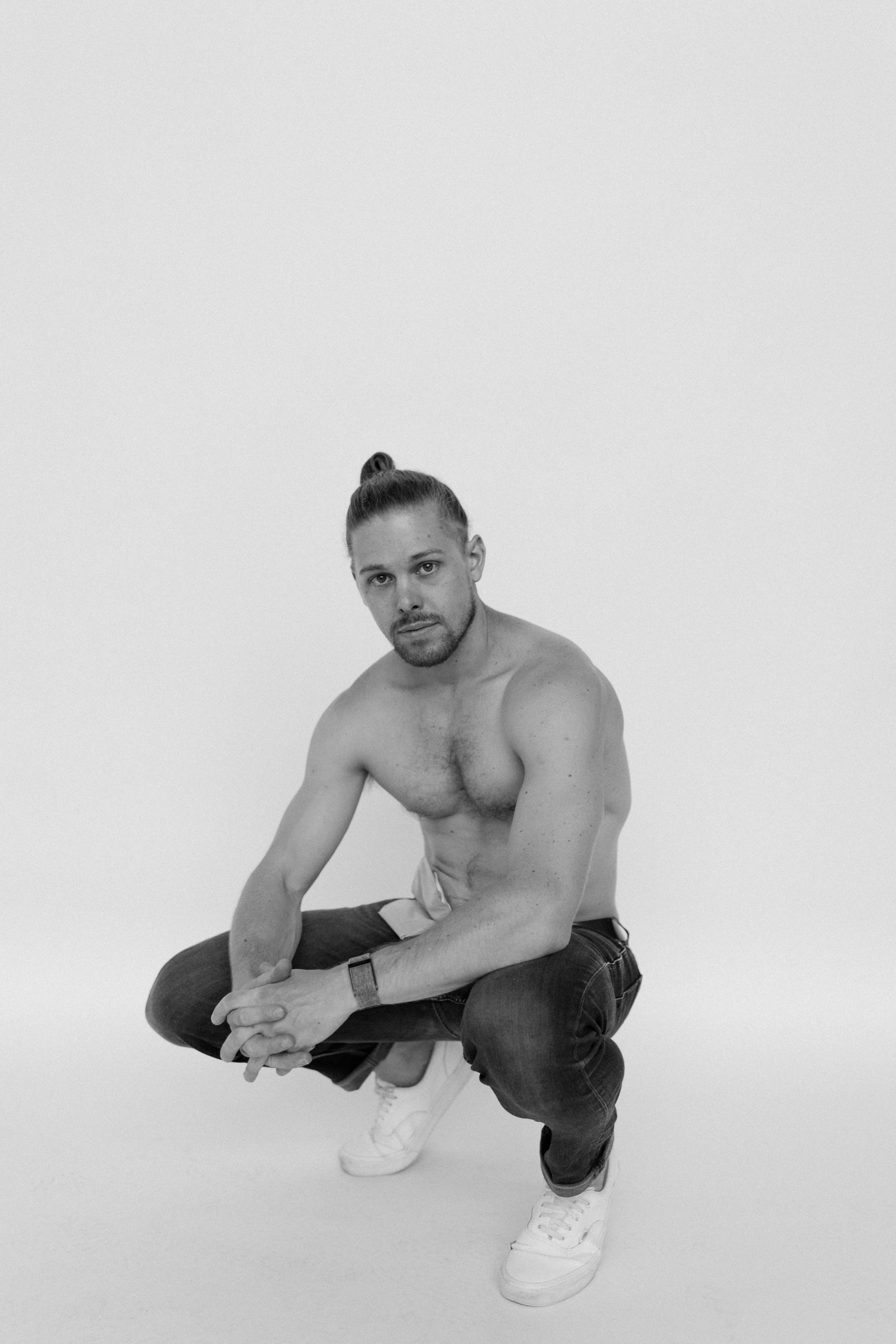 A shirtless man with a bun hairstyle crouching against a plain light background, wearing jeans, white sneakers, and a wristwatch.