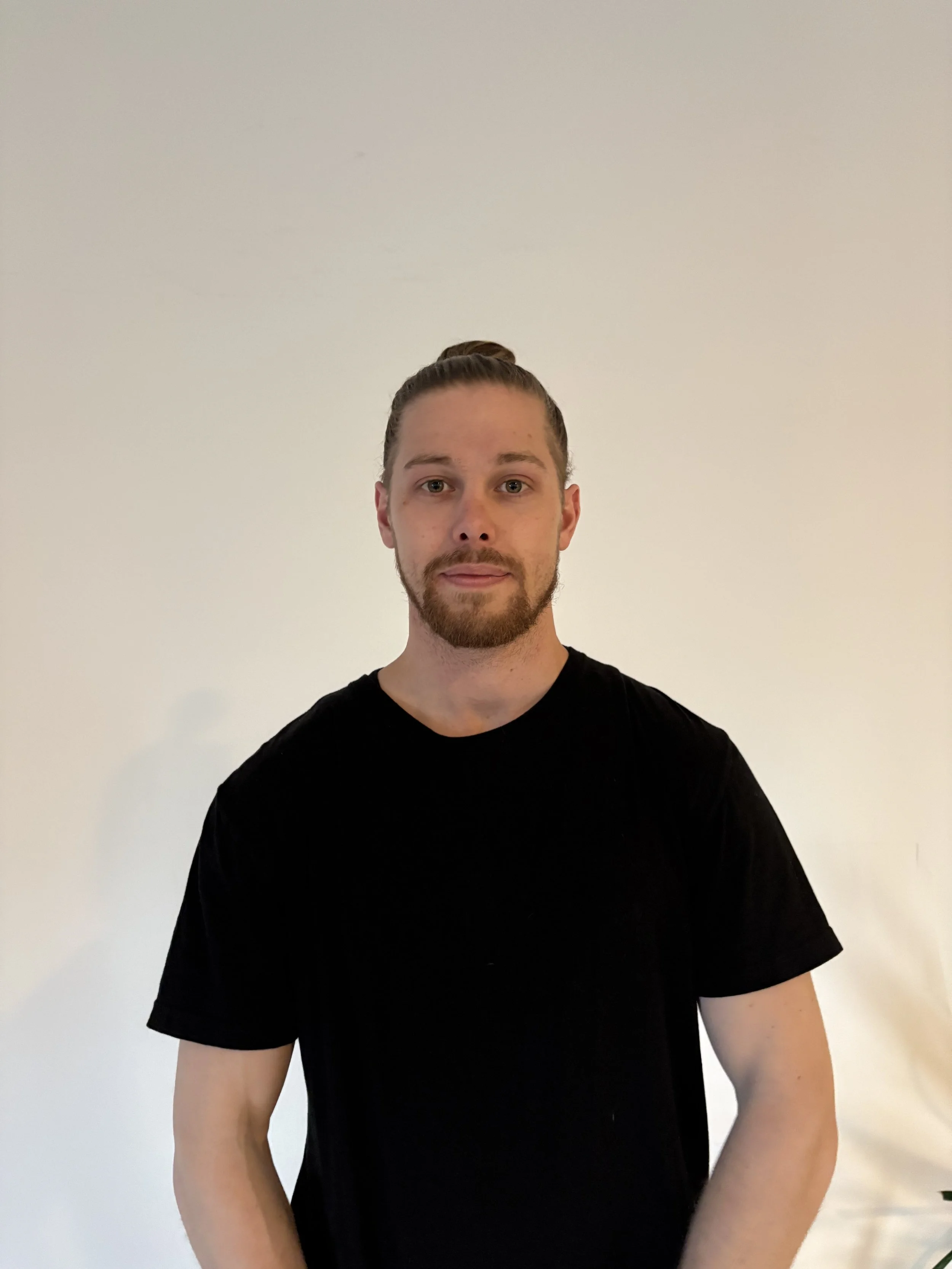 A man with a beard and ponytail wearing a black t-shirt standing against a plain white background.
